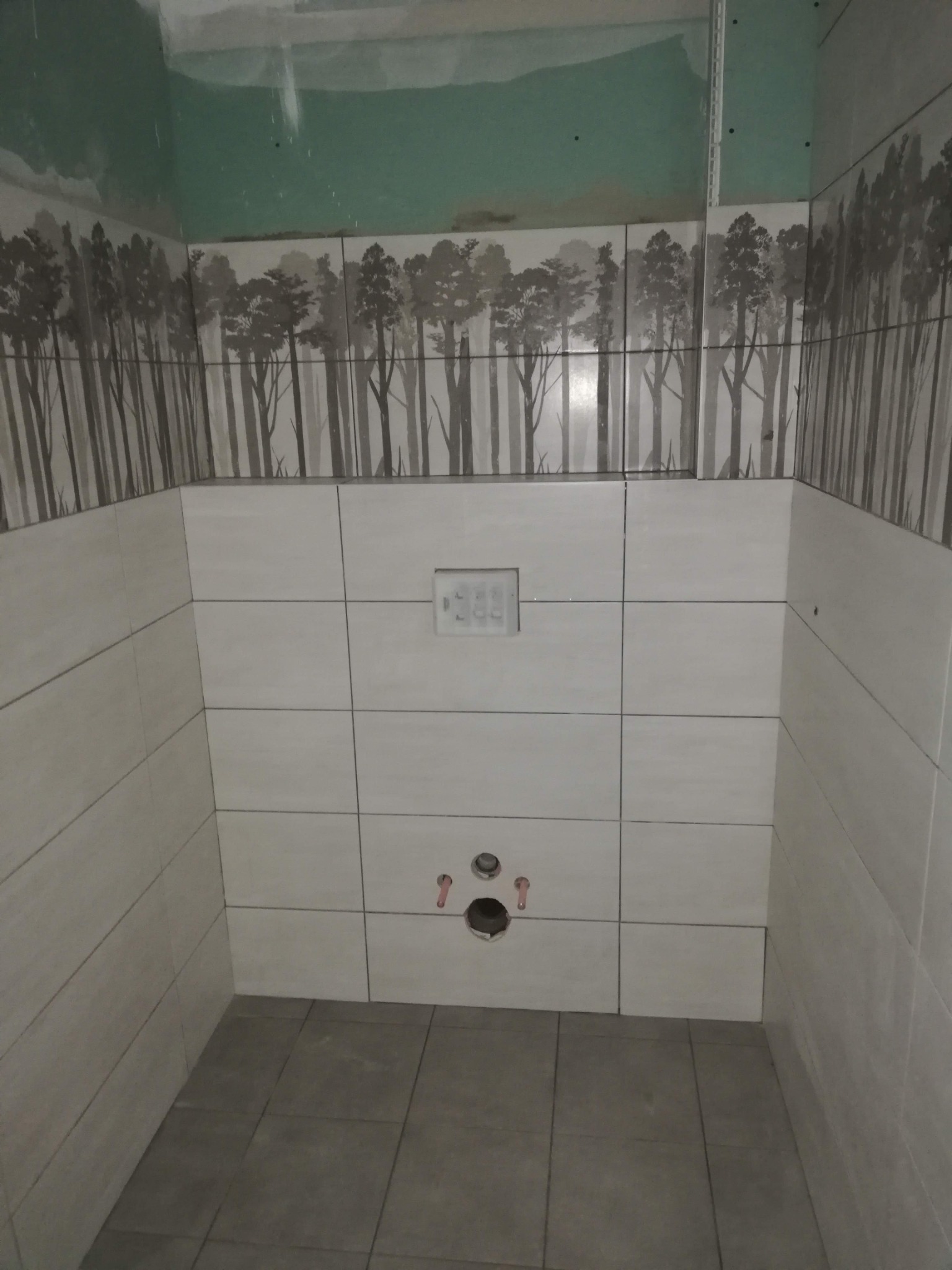 Wnętrze łazienki w trakcie remontu: ułożone jasne płytki z motywem lasu na górze, widoczne przyłącza wodne i częściowo wykończona ściana z zieloną farbą.