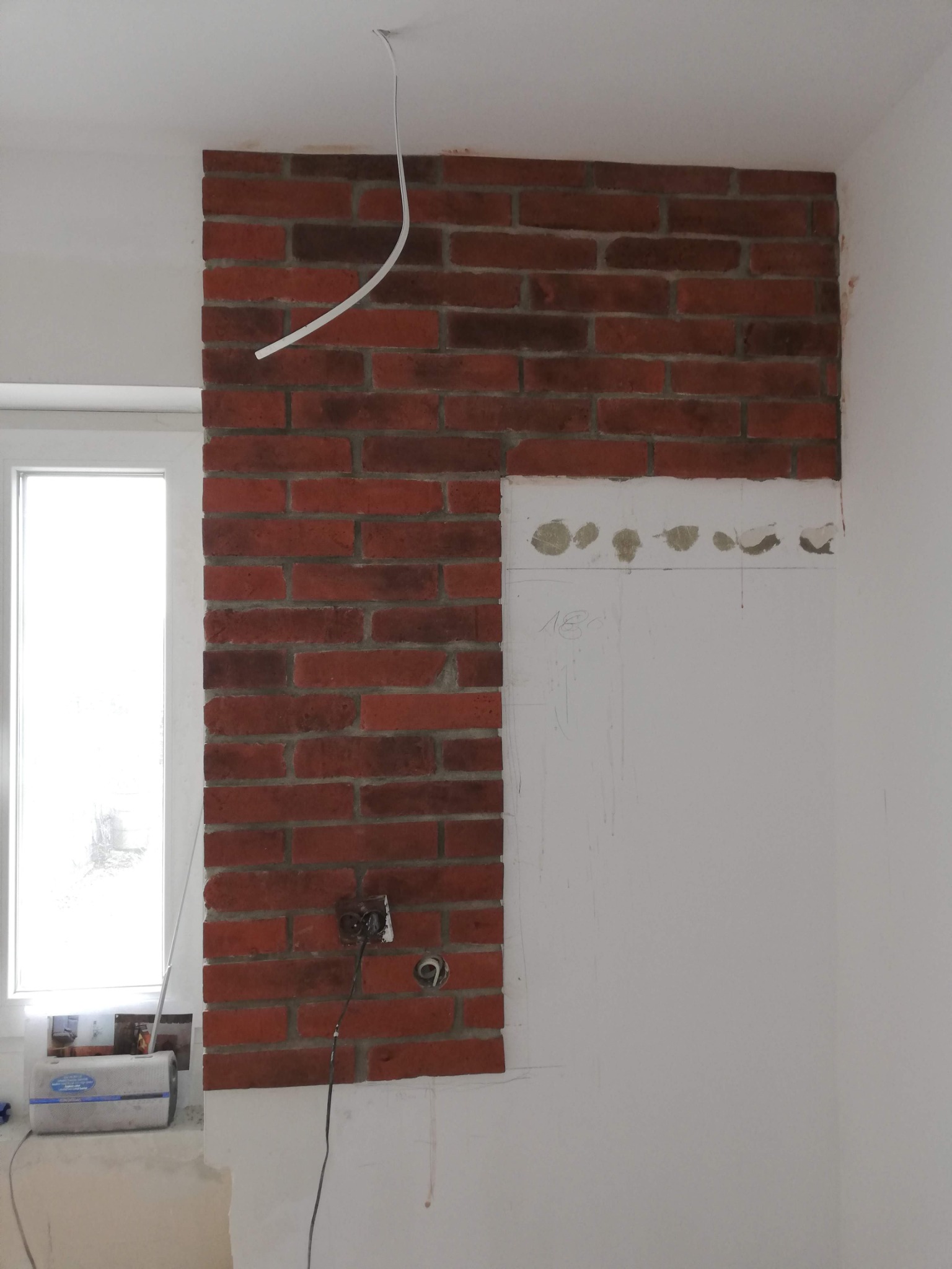 Fragment ściany z czerwoną cegłą klinkierową, częściowo położoną, z widocznym gniazdkiem elektrycznym i wystającym przewodem, obok okna i przygotowanej powierzchni ściany do dalszych prac...