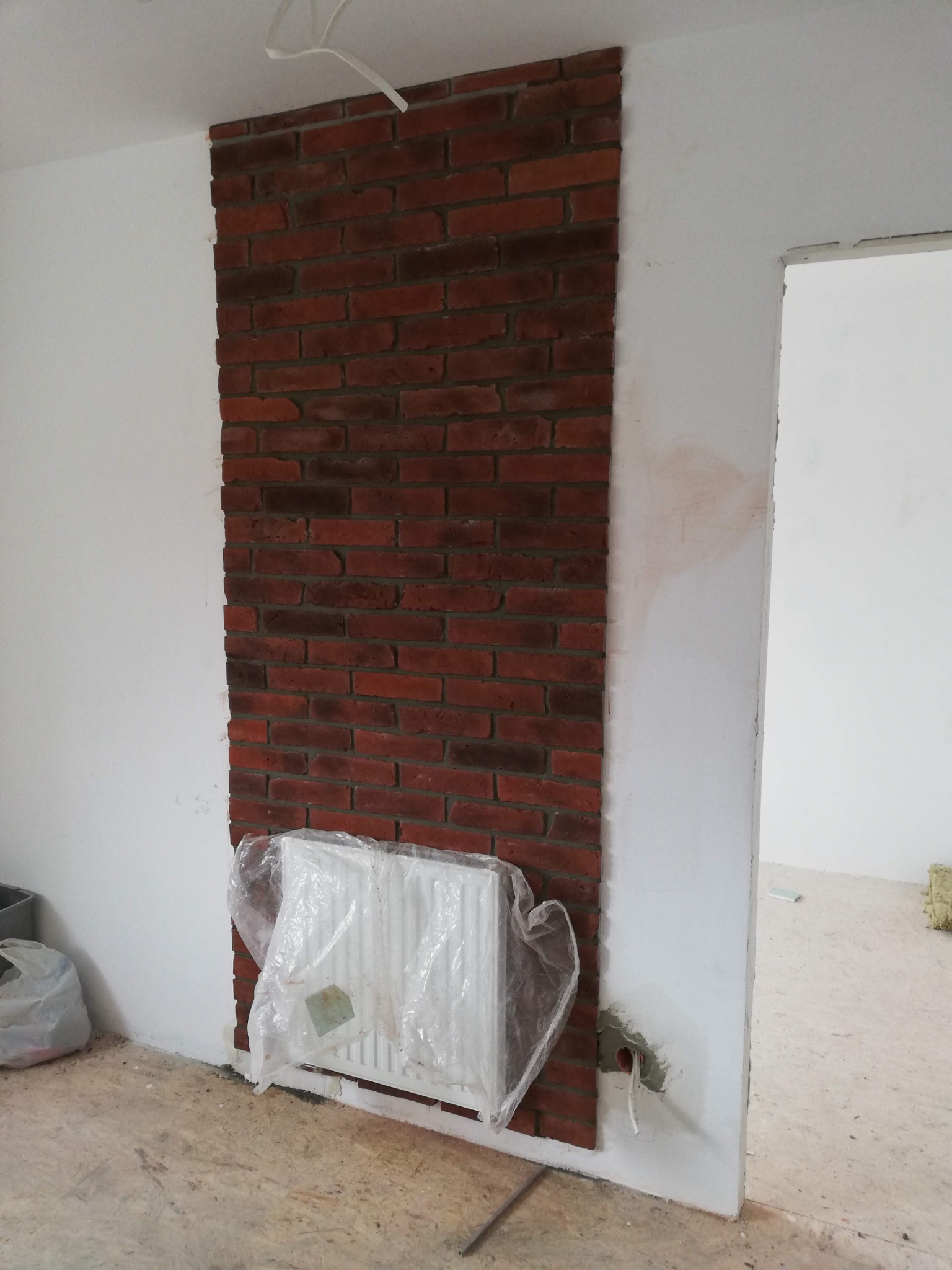 Fragment ściany z dekoracyjną okładziną z czerwonej cegły, zabezpieczony grzejnik folią, widoczne niedokończone prace wykończeniowe.