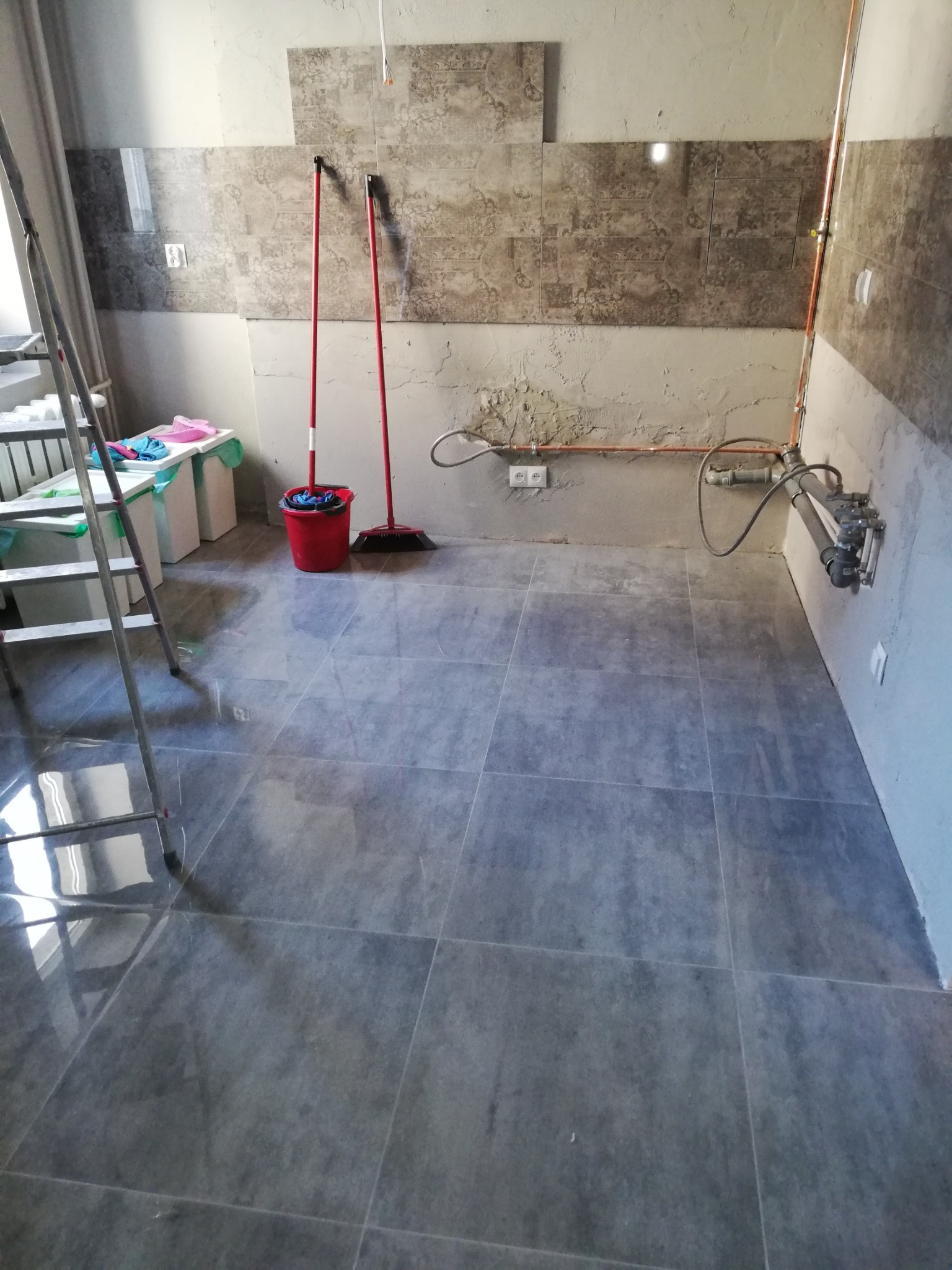 Szare płytki podłogowe ułożone w pomieszczeniu w trakcie remontu, widoczne fragmenty niedokończonej ściany z instalacją hydrauliczną i resztkami zaprawy, drabina i akcesoria malarskie po lewej...