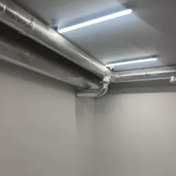 Izolowane rury wentylacyjne pod sufitem z oświetleniem LED i kamerą monitoringu w pomieszczeniu o białych ścianach i szarej podłodze.