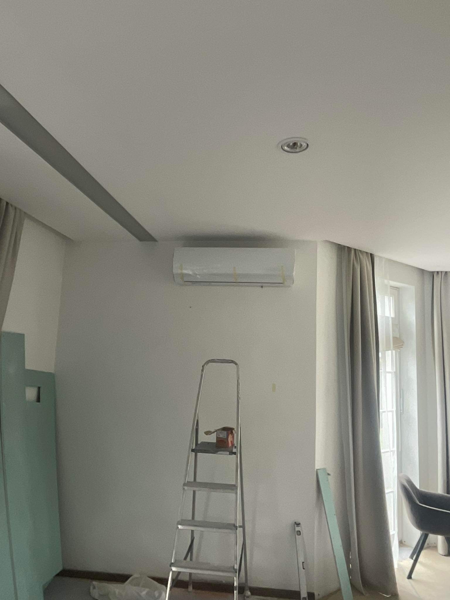 Montaż klimatyzatora w pokoju, widoczna drabina i zabezpieczony folią klimatyzator na ścianie, przygotowania do malowania ściany.