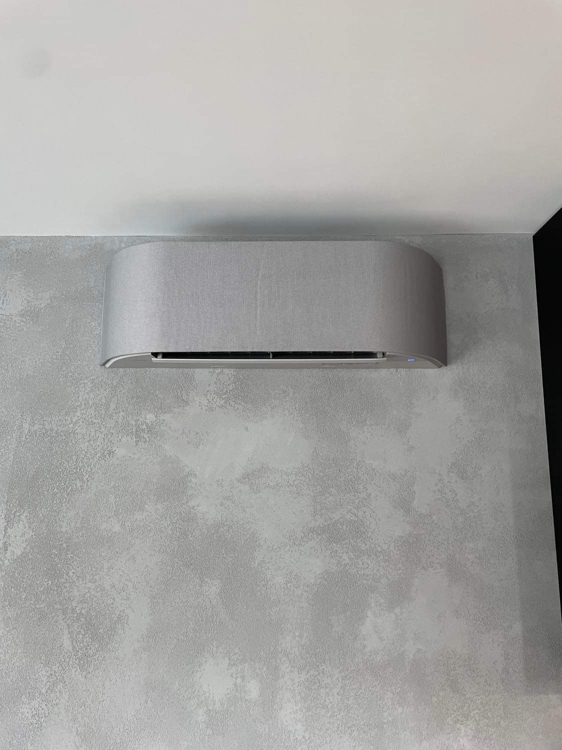 Nowoczesny klimatyzator w obudowie z szarej tkaniny zamontowany na ścianie o dekoracyjnej fakturze betonu.