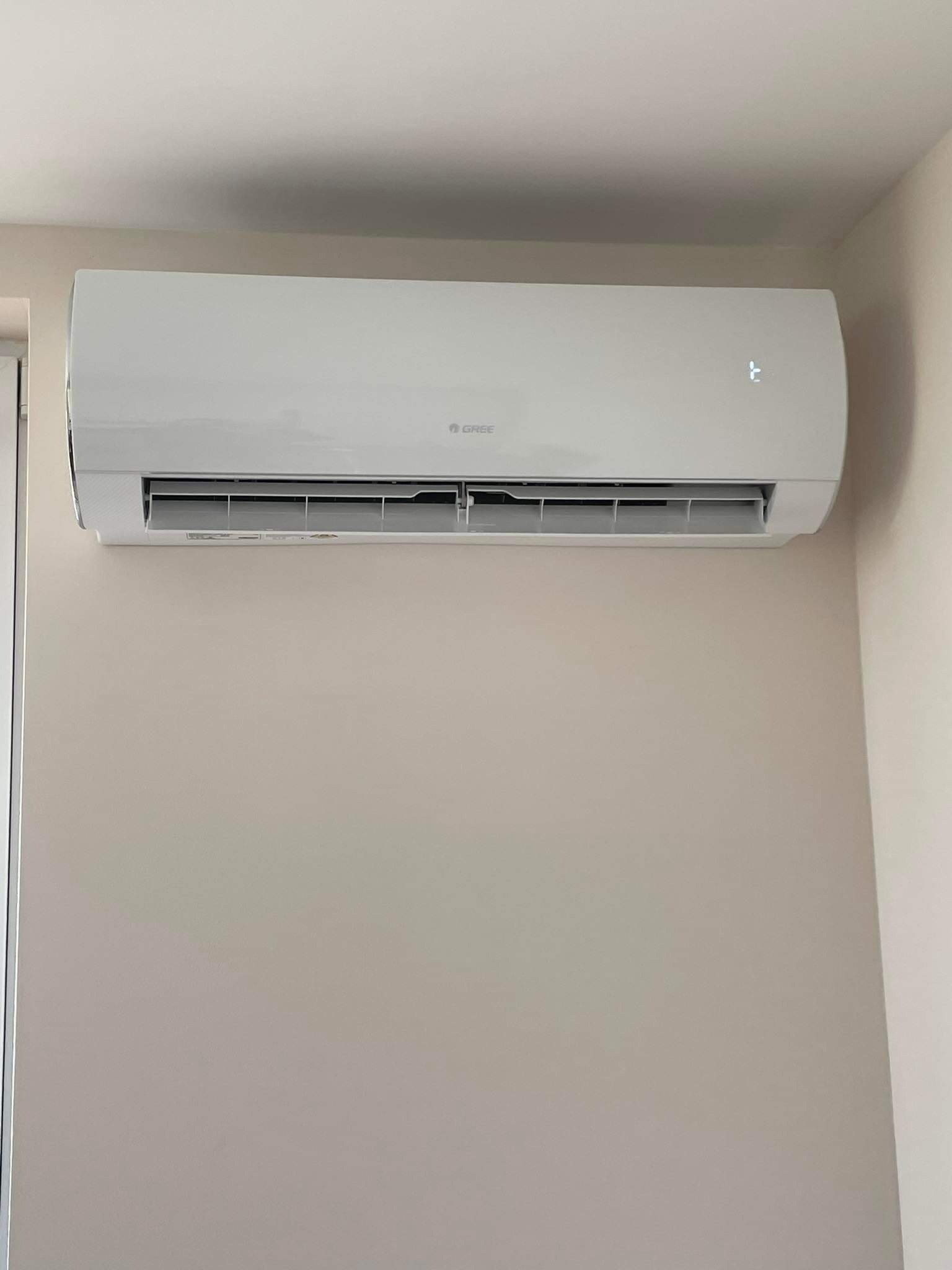 Biały klimatyzator marki Gree zamontowany na beżowej ścianie w pomieszczeniu, widoczne dyskretne cyfrowe wskazanie temperatury po prawej stronie urządzenia.