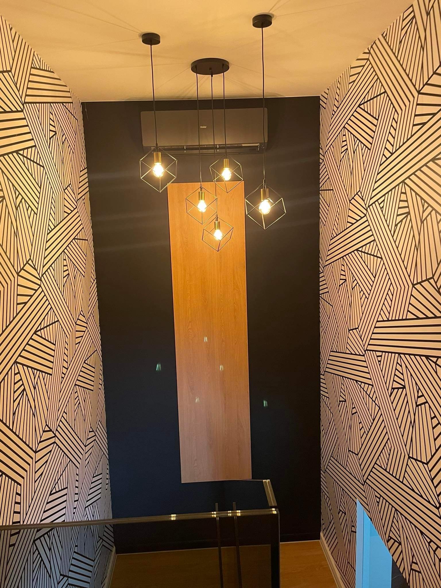 Wnętrze z klimatyzatorem Haier zamontowanym nad drewnianym panelem, oświetlone czterema lampami wiszącymi w geometrycznych kloszach, ściany pokryte tapetą w czarno-białe, abstrakcyjne wzory.