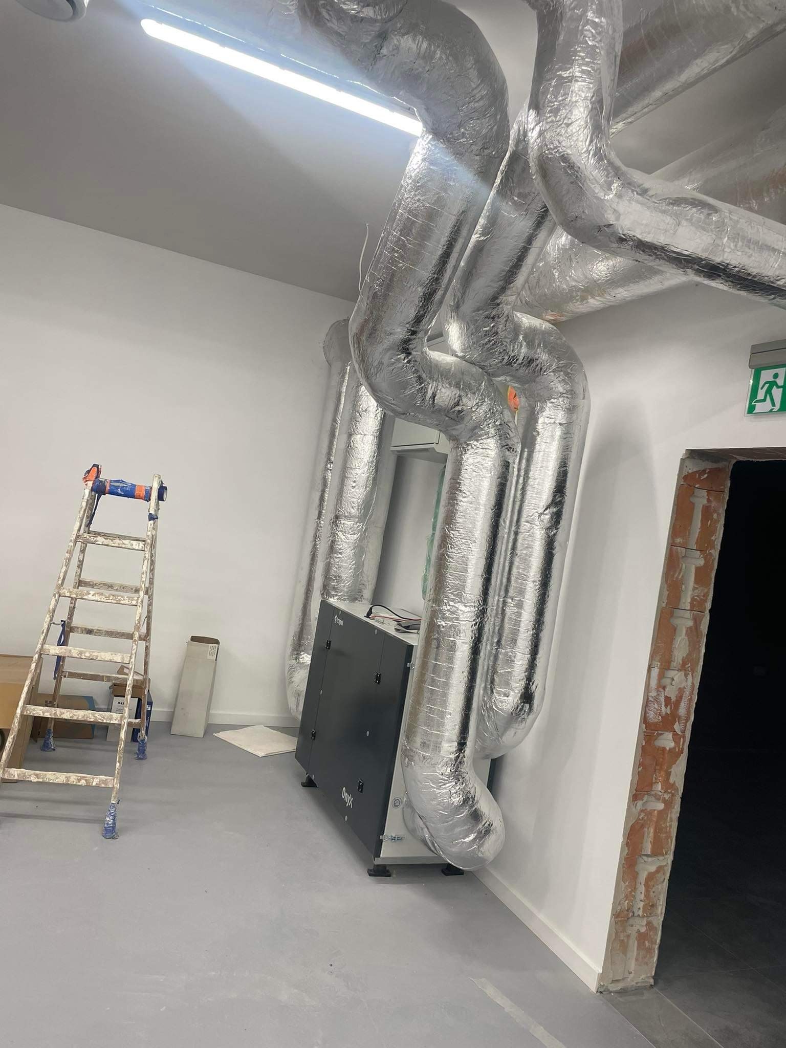 Montaż instalacji: srebrne, izolowane rury wentylacyjne prowadzone pod sufitem, obok drabiny malarskiej i wyjścia ewakuacyjnego w surowym wnętrzu.