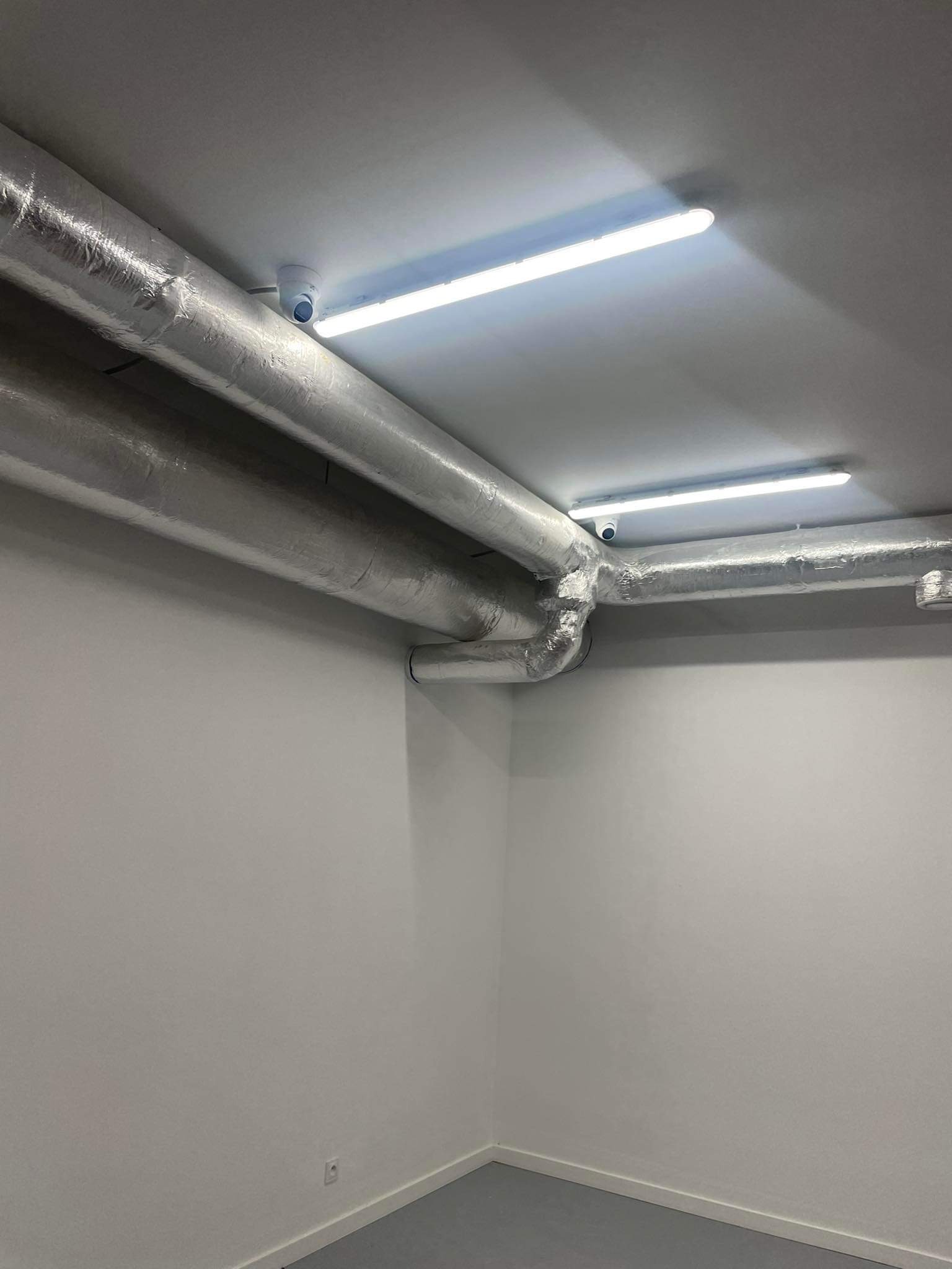 Izolowane rury wentylacyjne pod sufitem z oświetleniem LED i kamerą monitoringu w pomieszczeniu o białych ścianach i szarej podłodze.