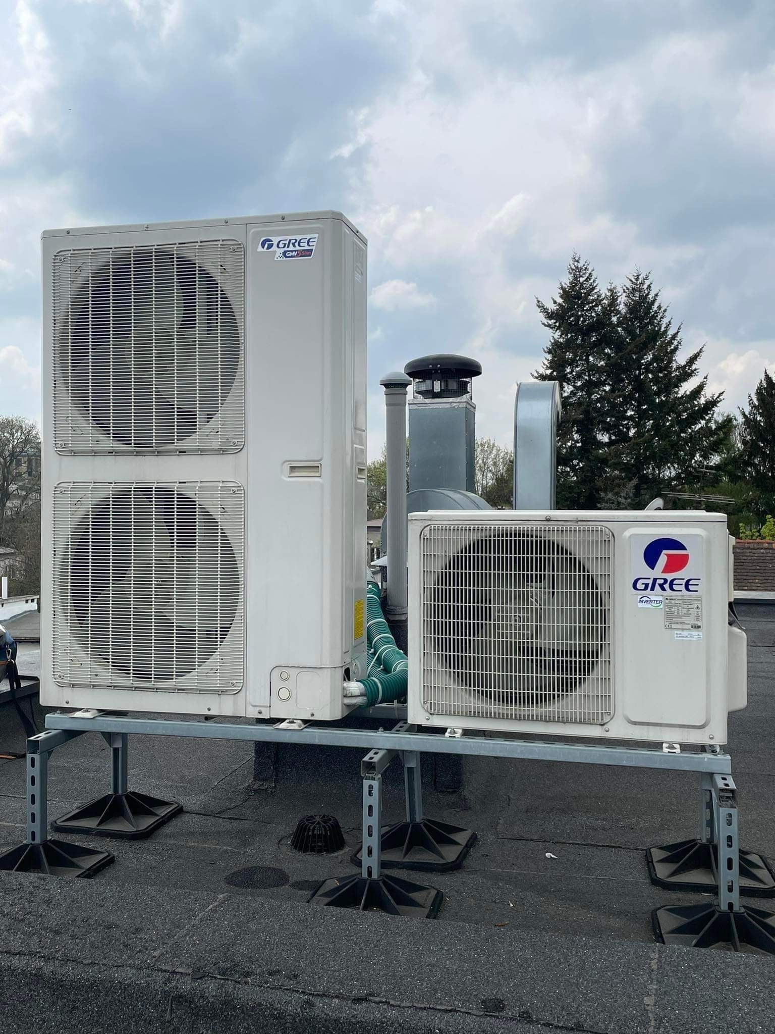 Dwa agregaty klimatyzacji marki Gree na dachu pokrytym papą, zamontowane na metalowej konstrukcji wsporczej z gumowymi podkładkami antywibracyjnymi, widoczne kominy wentylacyjne w tle i pochmurne...