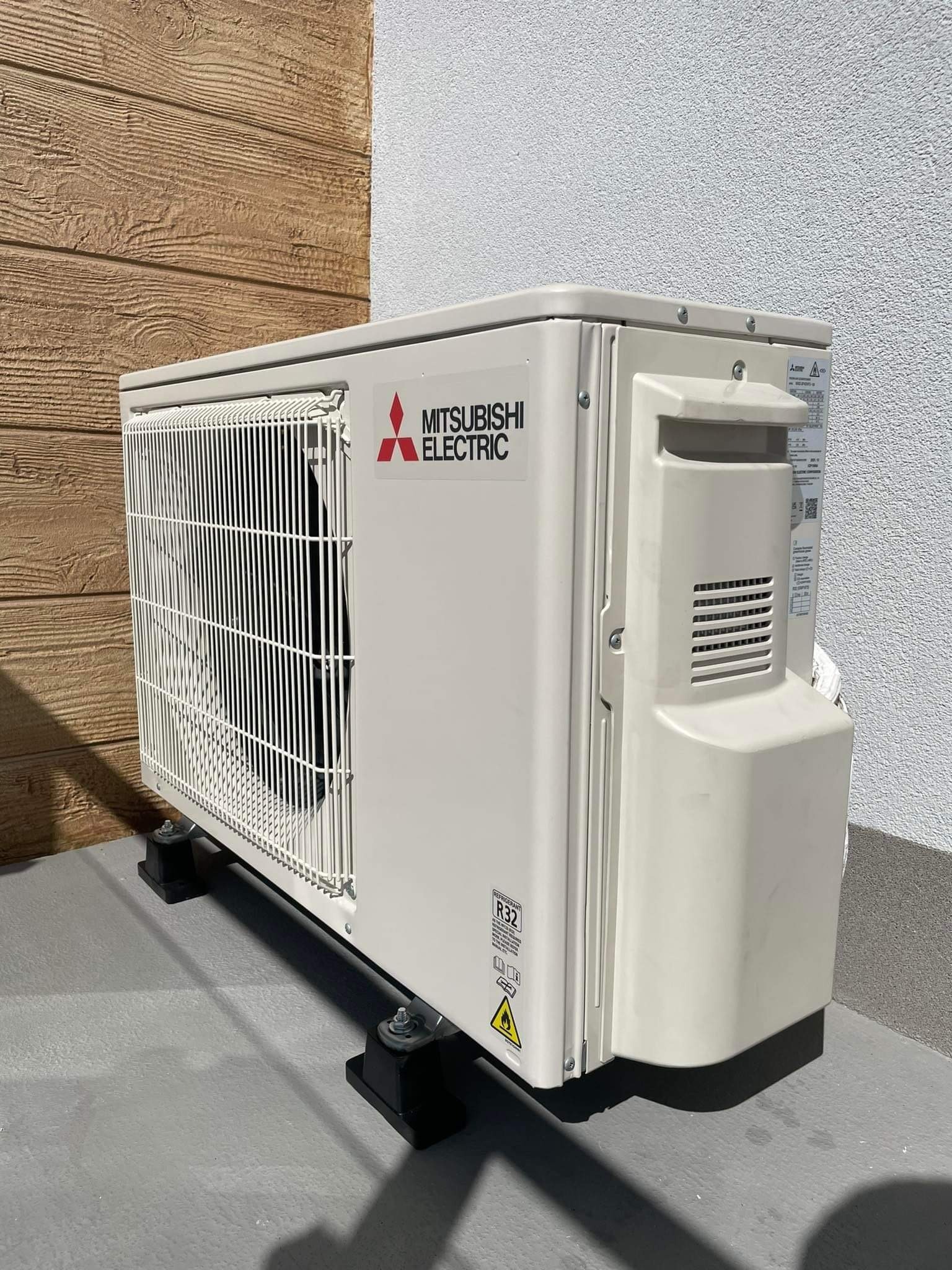 Zewnętrzna jednostka klimatyzacji Mitsubishi Electric zamontowana na czarnych podstawkach, widoczna naklejka z symbolem R32, na tle ściany z fakturą drewna i białego tynku.
