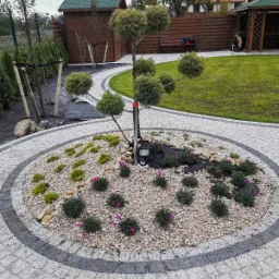 Okrągły klomb z ozdobnym drzewkiem formowanym w kule, wysypany białym żwirem, z nasadzeniami mchu i niskich roślin kwitnących na różowo, otoczony kostką brukową, z oświetleniem punktowym.