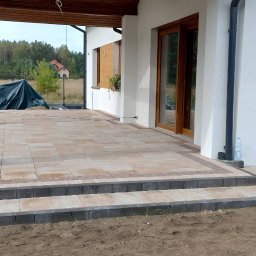 Jaworex - Wykończony taras z kostki brukowej w odcieniach beżu i brązu, prowadzący do domu z białymi ścianami i drewnianymi oknami, widok na ogród w tle, świeżo posadzone rośliny w donicach...