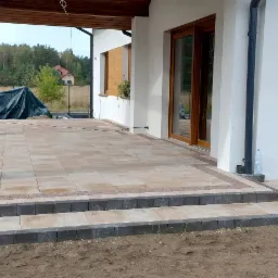 Wykończony taras z kostki brukowej w odcieniach beżu i brązu, prowadzący do domu z białymi ścianami i drewnianymi oknami, widok na ogród w tle, świeżo posadzone rośliny w donicach...