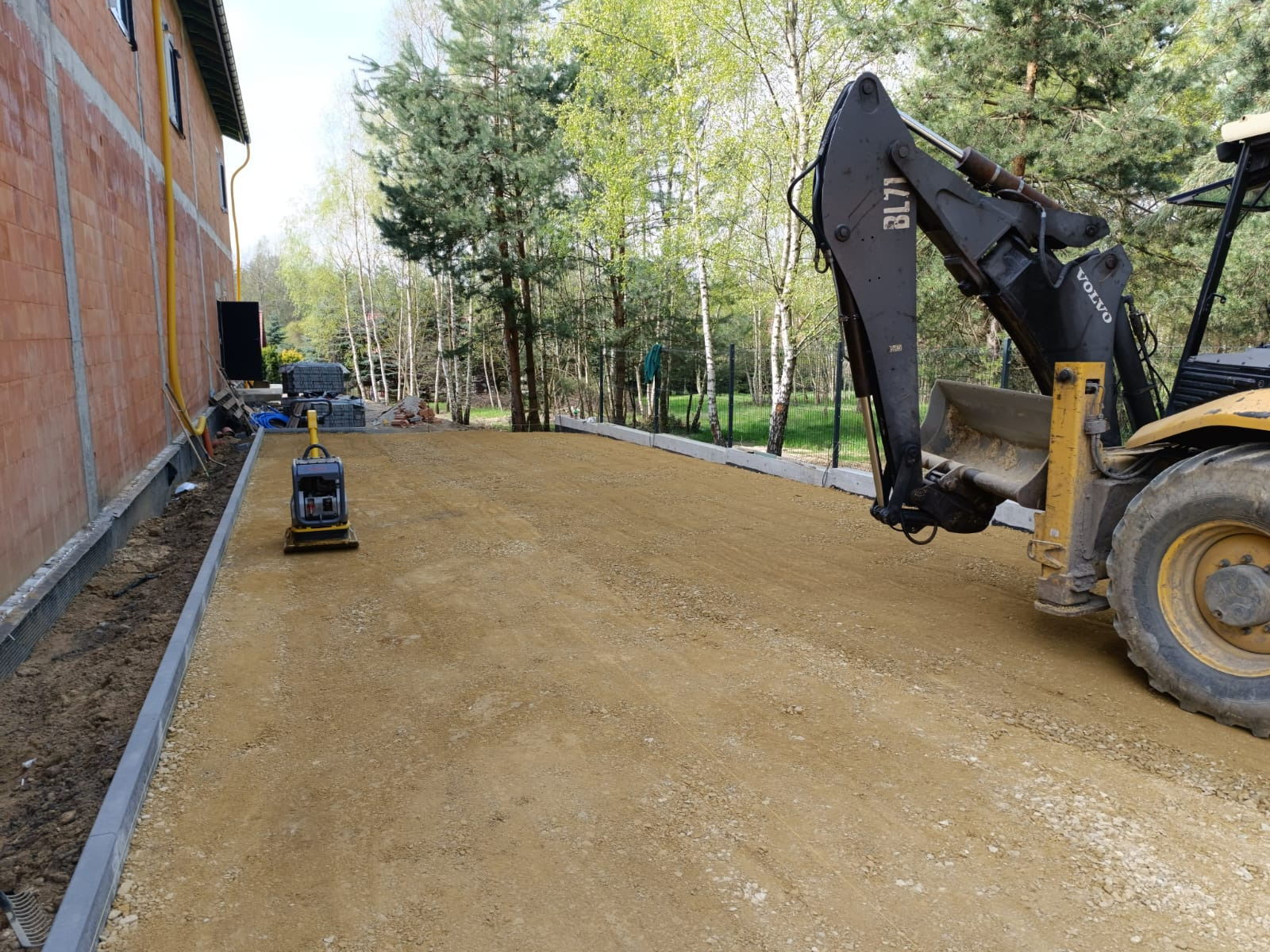 Przygotowanie podłoża pod nawierzchnię z użyciem zagęszczarki i koparko-ładowarki Volvo BL71, widoczne obrzeża betonowe i podsypka piaskowa