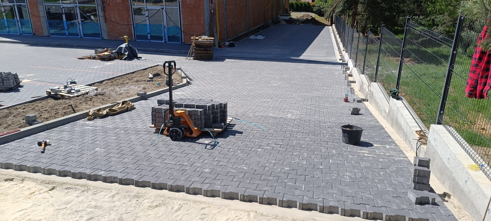 Układanie szarej kostki brukowej typu 'I' na posesji z ogrodzeniem panelowym, widoczny paleciak z kostką, gumowy młotek i czarne wiadro, w tle budynek w stanie surowym.