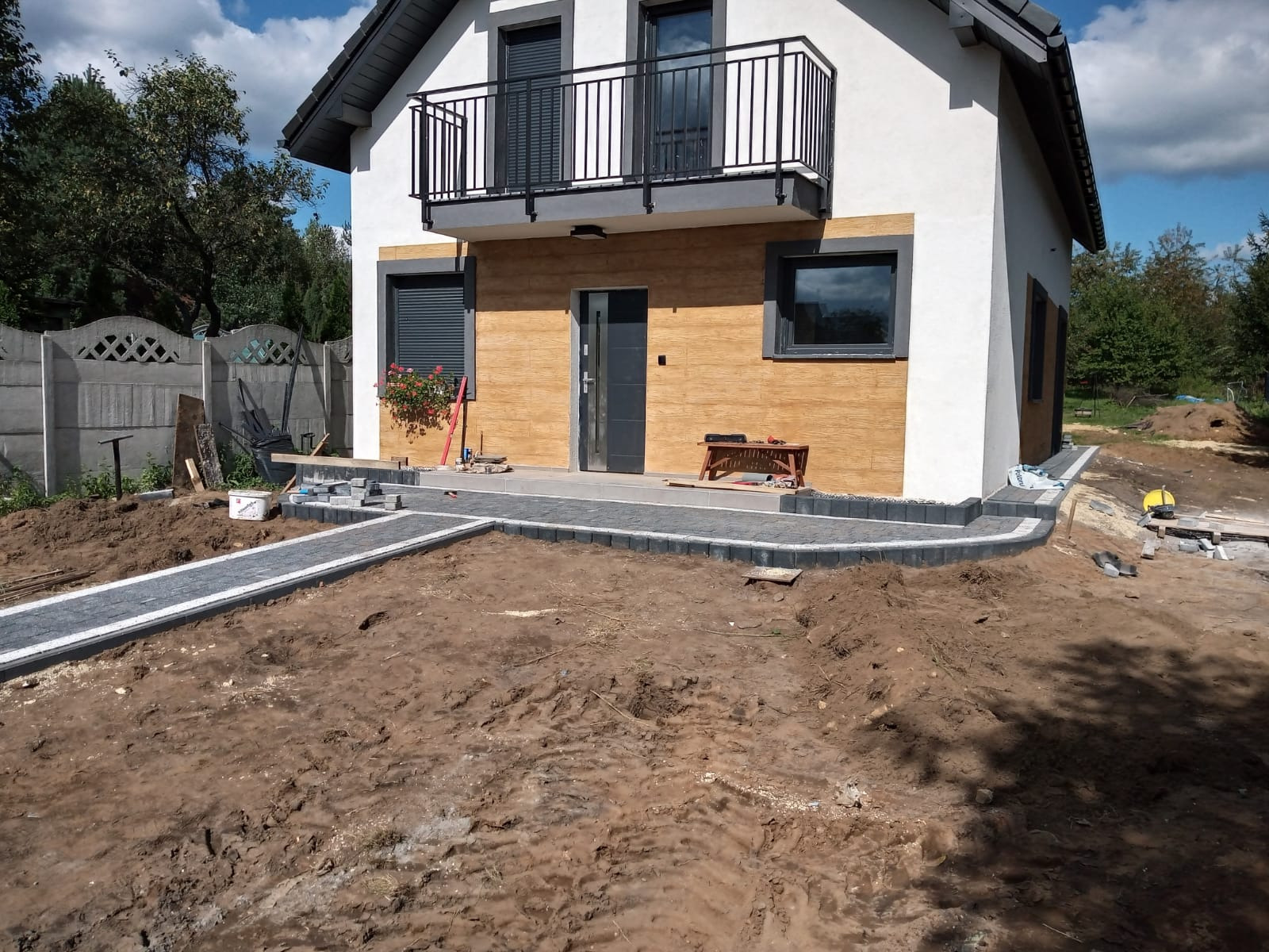 Nowoczesny dom z jasną elewacją i drewnianą okładziną, widoczny fragment ścieżki z szarych kostek z białymi obrzeżami w trakcie budowy, ziemia wokół przygotowana pod dalsze prace.