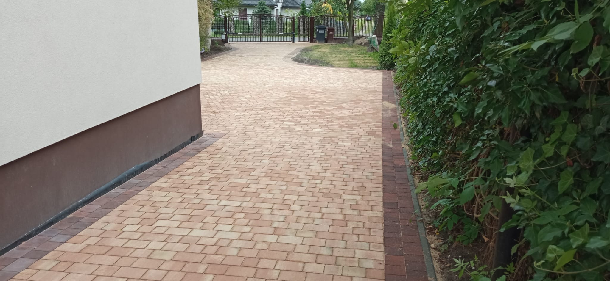 Ułożona kostka brukowa w odcieniach beżu i brązu tworząca podjazd do domu, obrzeże z ciemniejszej kostki, widoczna ściana budynku i żywopłot.