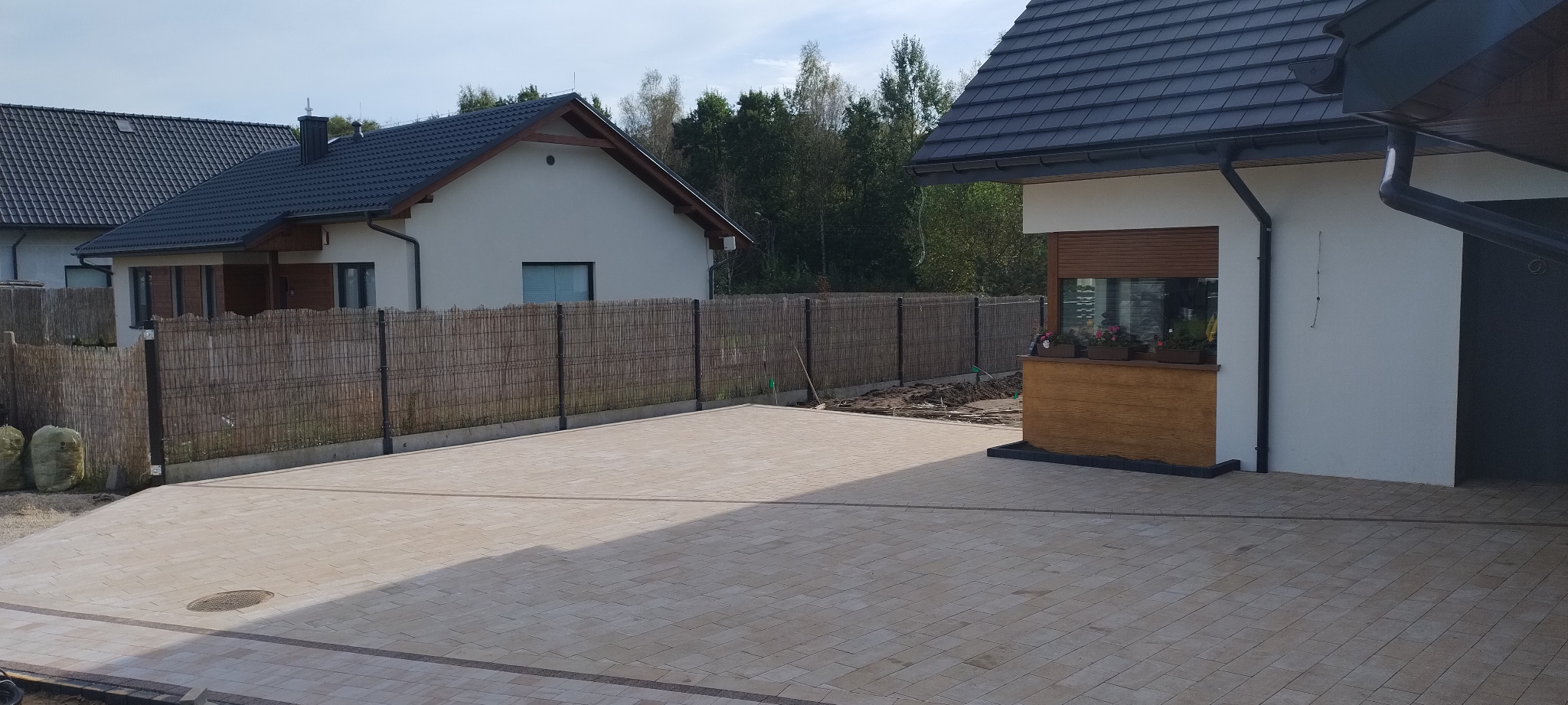 Ułożona beżowa kostka brukowa na podjeździe przed domem z bambusowym ogrodzeniem i rabatą kwiatową, widoczny fragment dachu drugiego budynku.