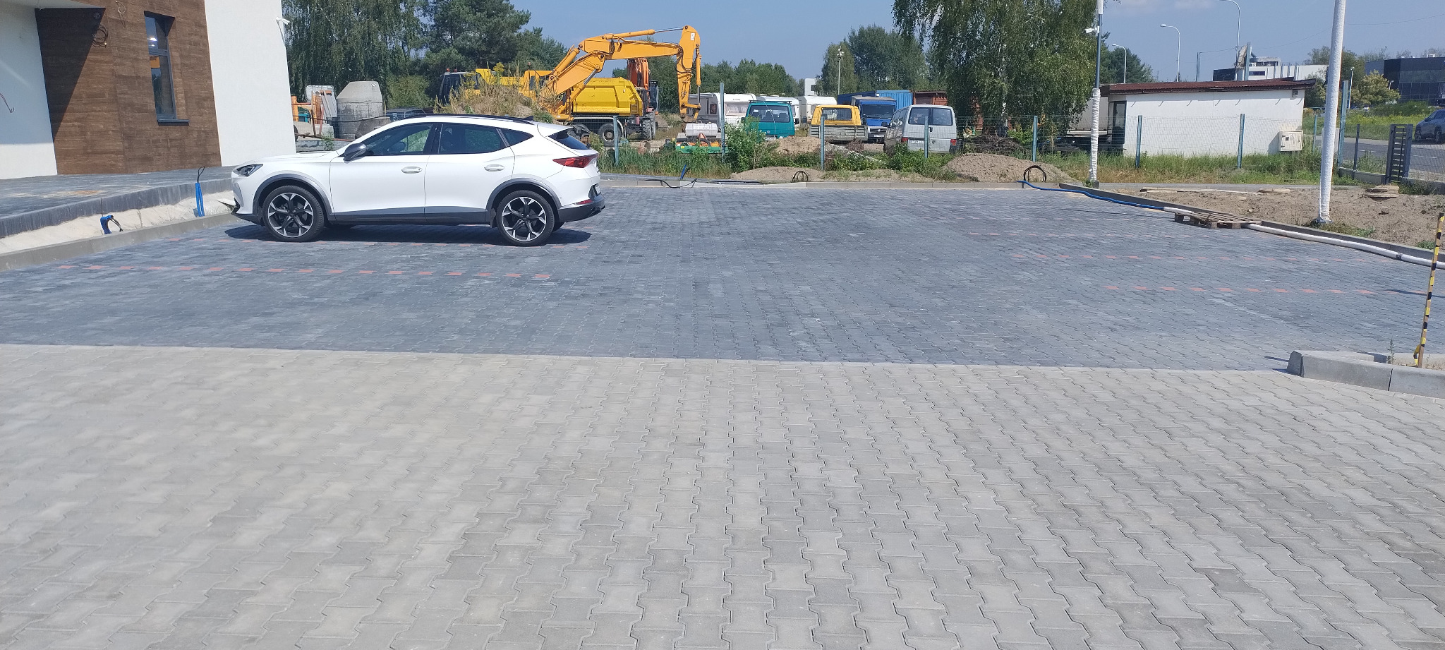 Nowo ułożona, dwukolorowa kostka brukowa na parkingu z zaparkowanym białym samochodem i sprzętem budowlanym w tle.