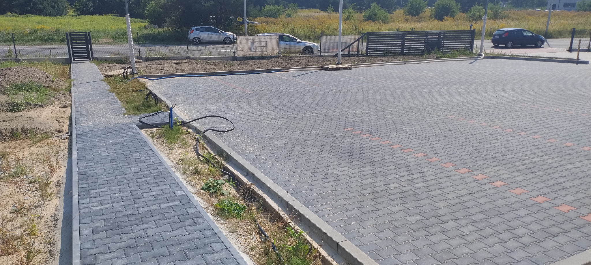 Ukończony parking z szarej kostki brukowej z czerwonymi akcentami, widoczny chodnik z kostki prowadzący do bramy, w tle zaparkowane samochody i zieleń.