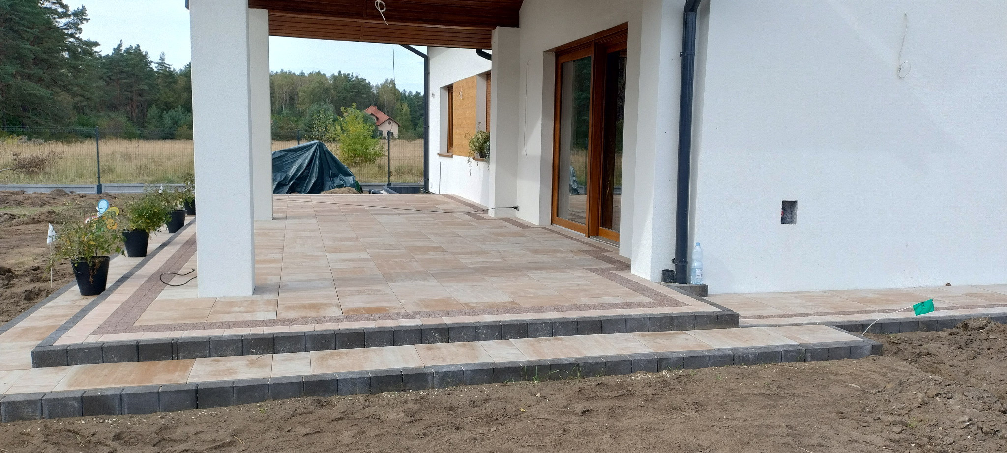 Wykończony taras z kostki brukowej w odcieniach beżu i brązu, prowadzący do domu z białymi ścianami i drewnianymi oknami, widok na ogród w tle, świeżo posadzone rośliny w donicach...