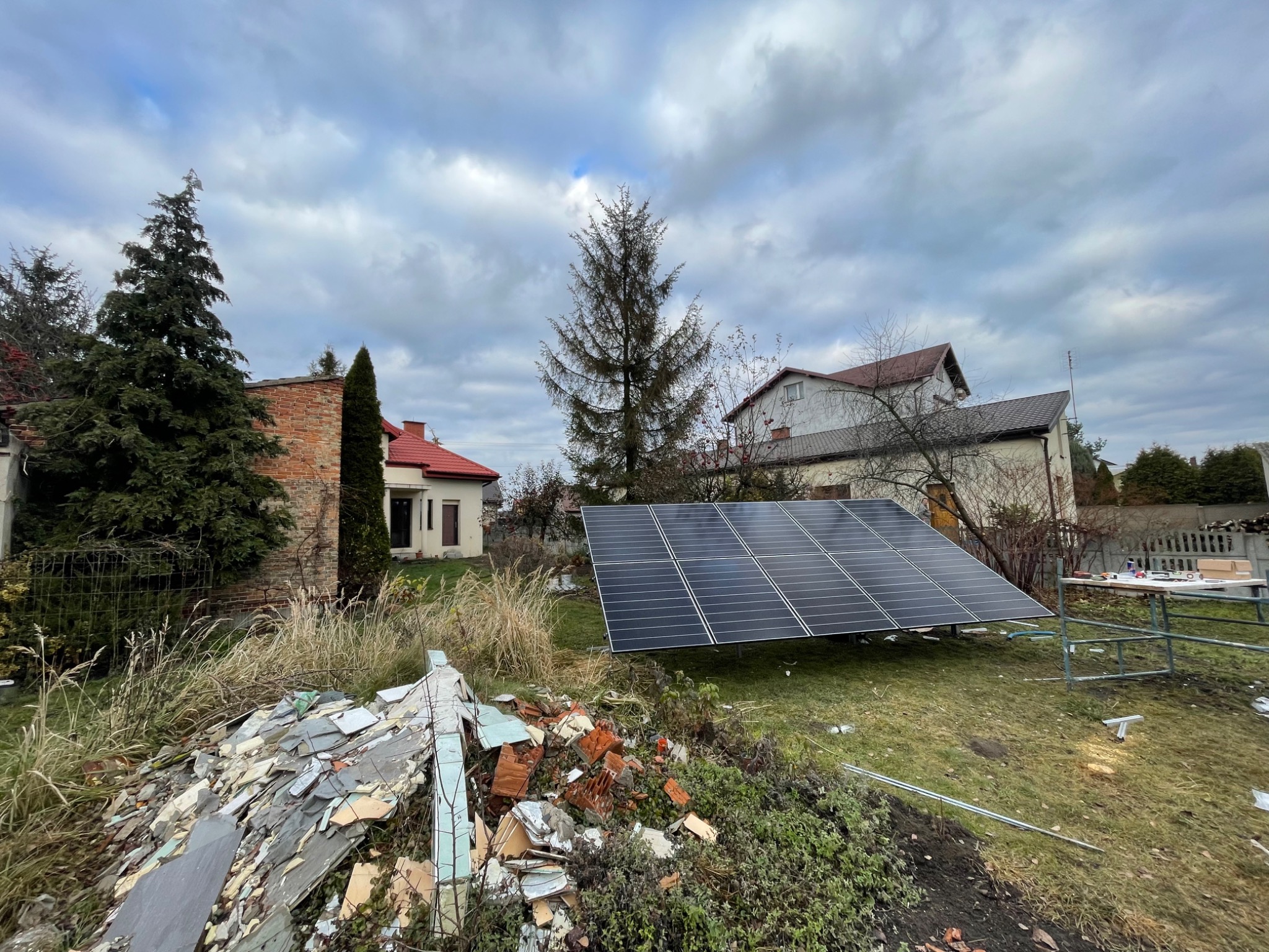 Panele słoneczne ustawione na trawniku przed domem, z gruzem w rogu kadru i stołem montażowym w tle, pochmurne niebo.