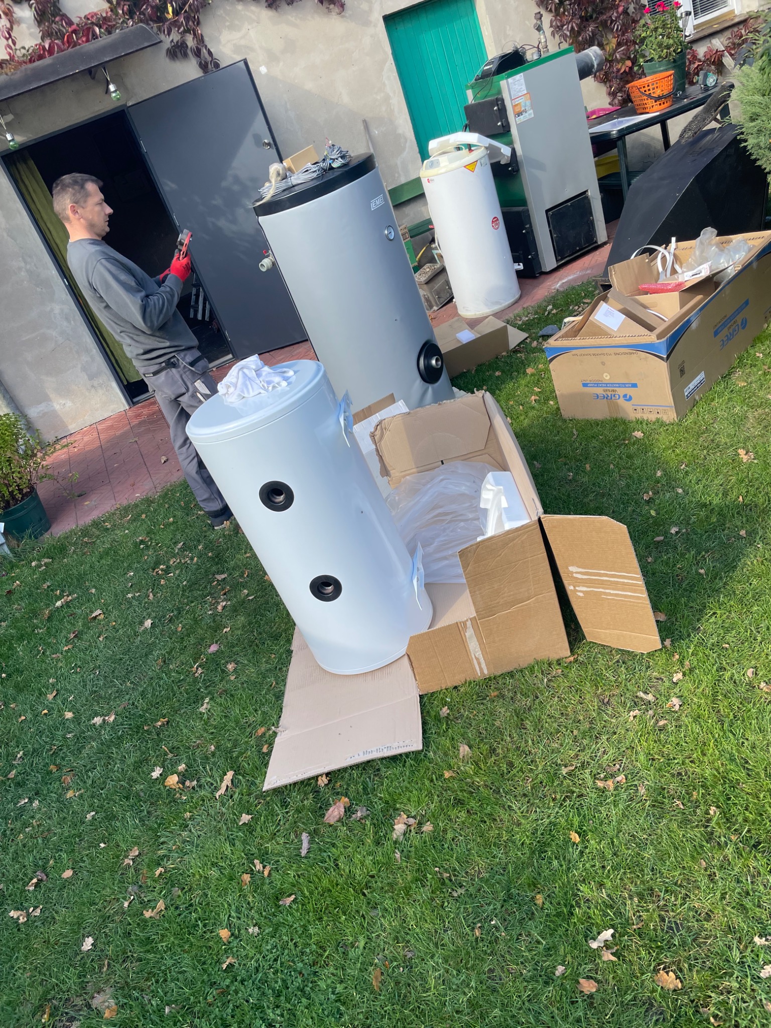 Instalator w czerwonych rękawicach przygotowuje biały zasobnik ciepłej wody użytkowej na trawniku przed budynkiem, obok rozpakowane kartony i inne elementy instalacji.