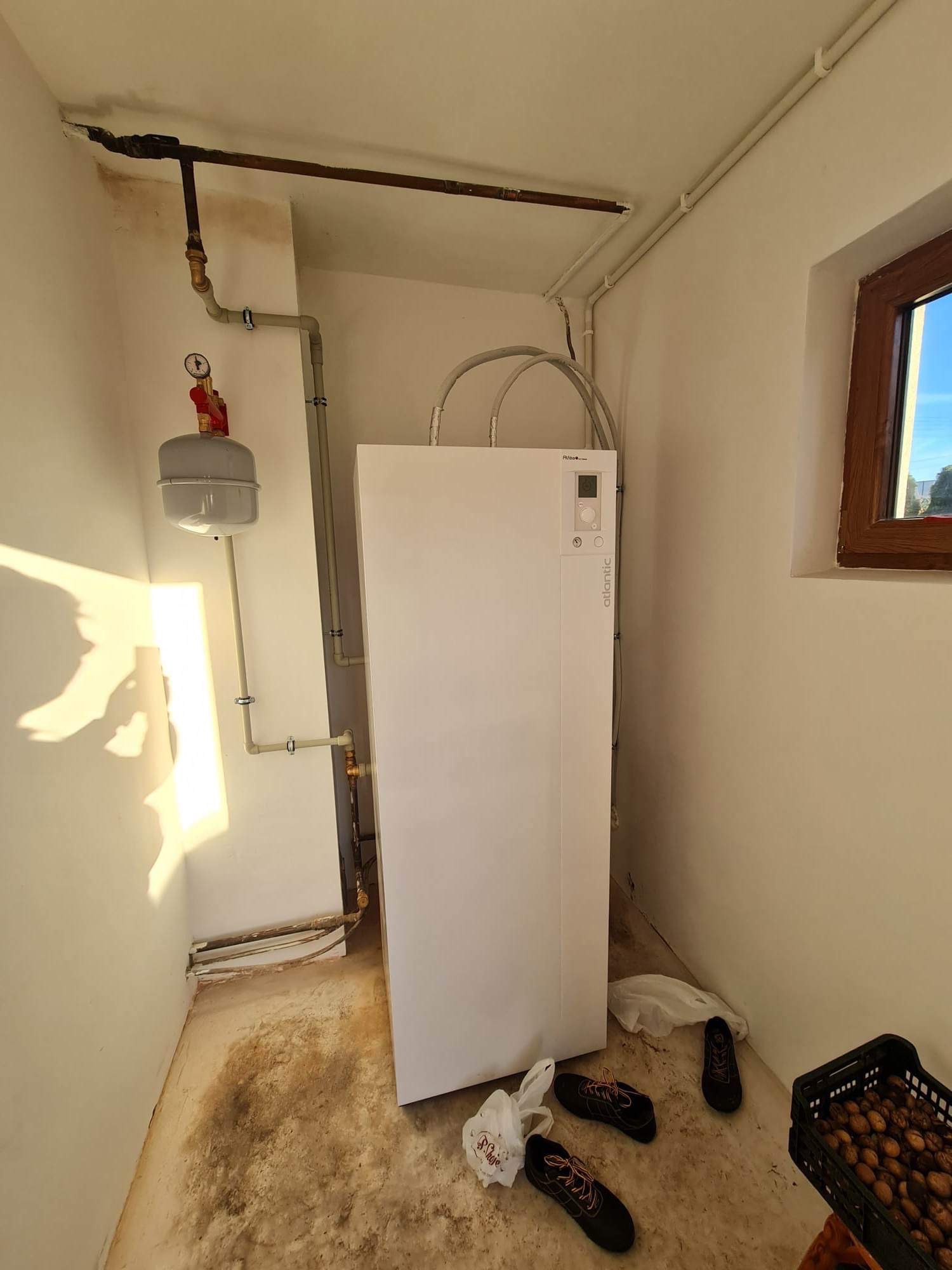 Biała pompa ciepła Atlantic zamontowana w małym pomieszczeniu z instalacją hydrauliczną, widoczne rury, zbiornik wyrównawczy, okno i akcesoria na podłodze.