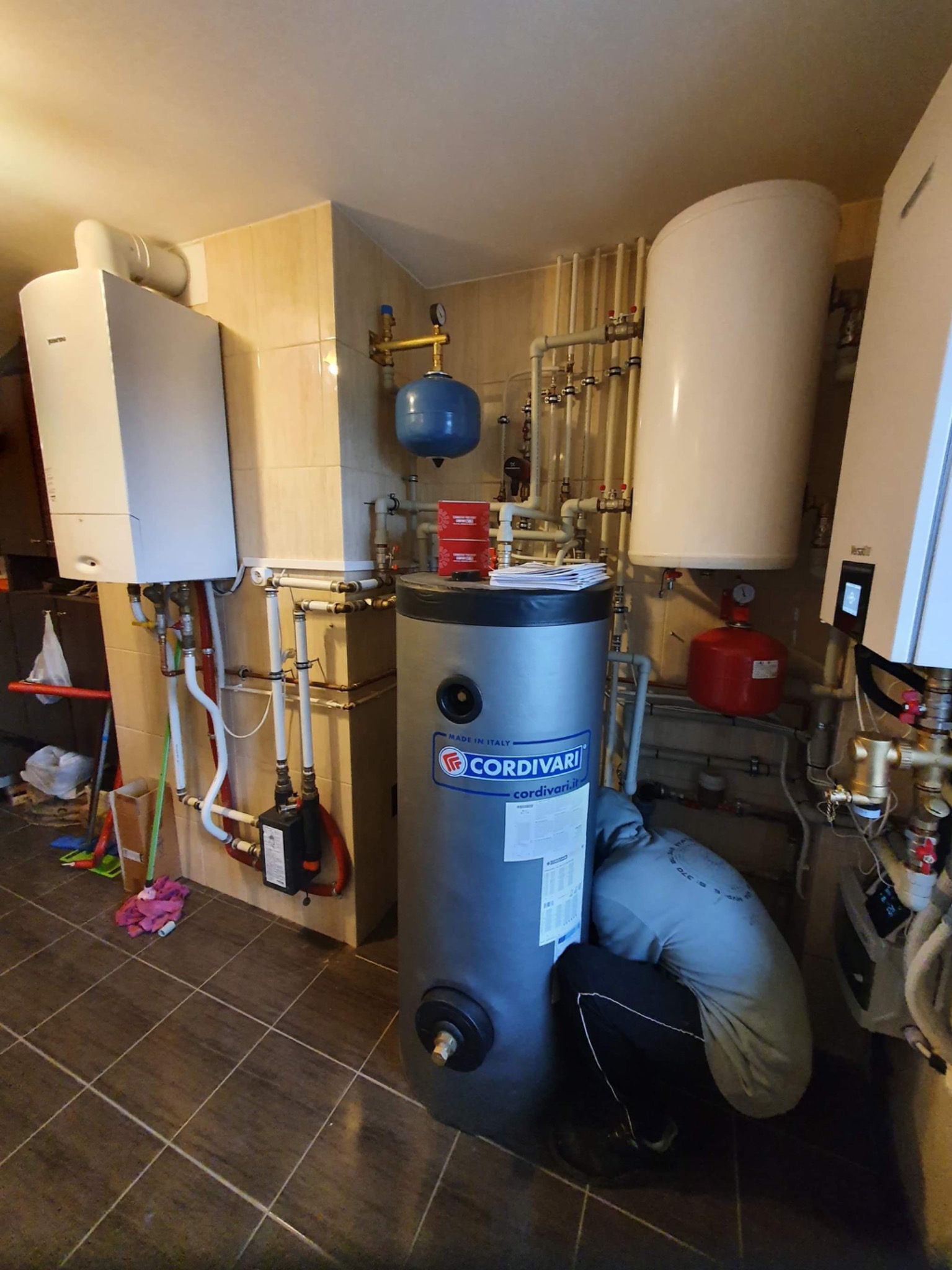 Instalacja centralnego ogrzewania w pomieszczeniu z kotłem gazowym, zasobnikiem ciepłej wody Cordivari i licznymi rurami oraz osprzętem, osoba w trakcie pracy przy zasobniku.