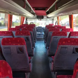 Wnętrze autobusu z czerwonymi siedzeniami z geometrycznym wzorem, widok na kokpit kierowcy i krajobraz za oknem z widocznym znakiem drogowym.