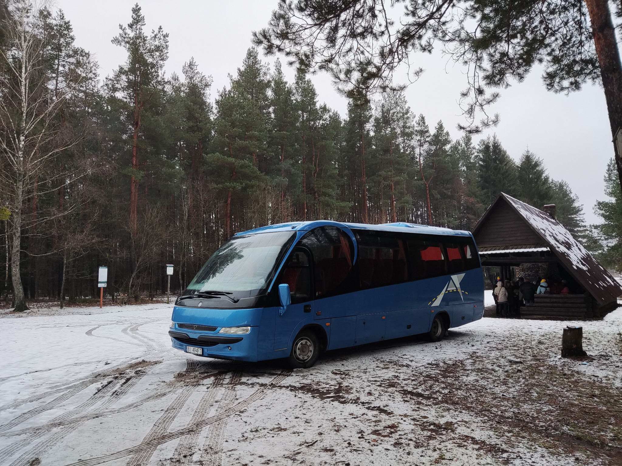 Niebieski autobus turystyczny zaparkowany na ośnieżonym parkingu leśnym obok drewnianej wiaty z grupą osób wewnątrz.