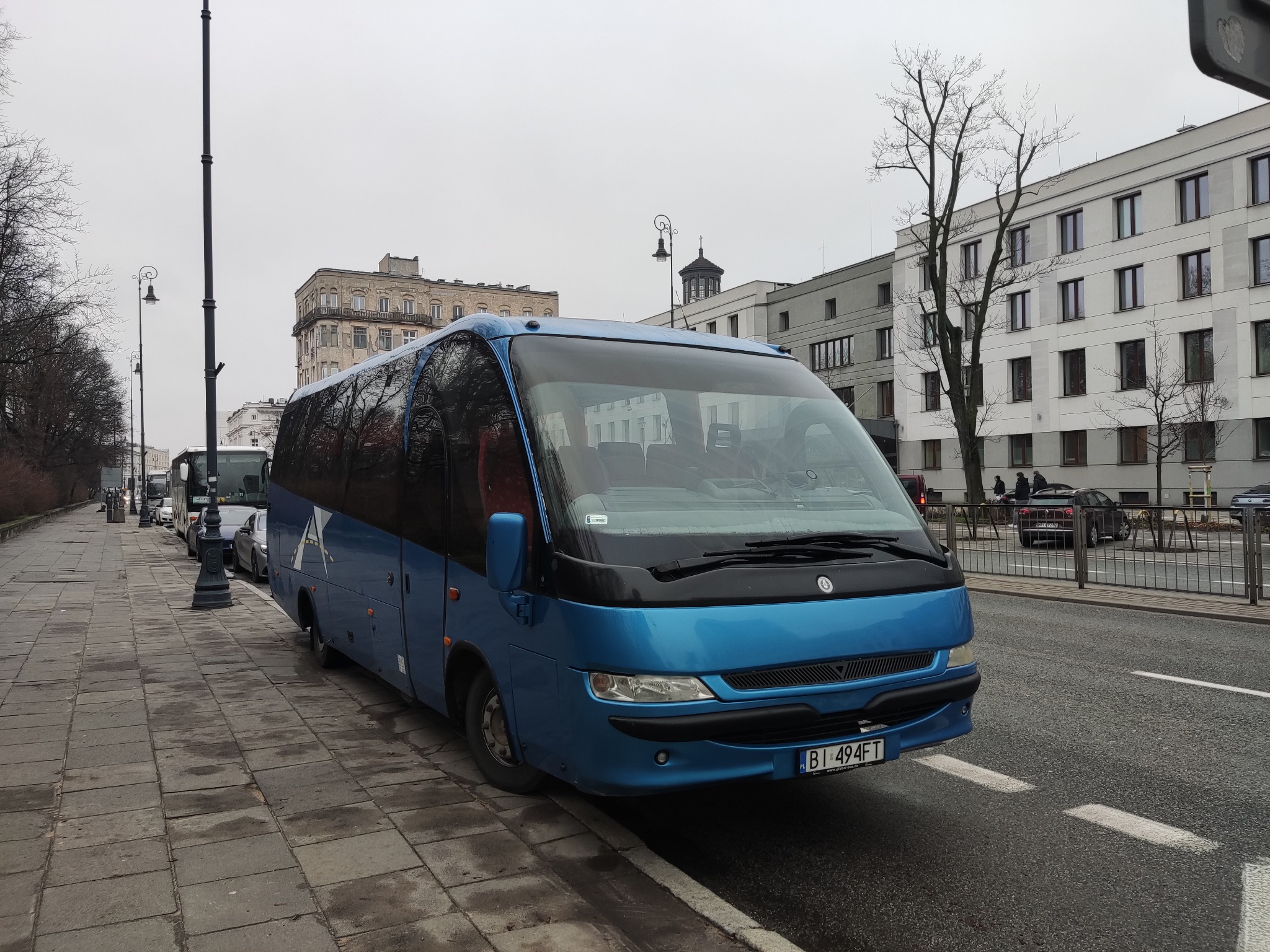 Niebieski bus turystyczny zaparkowany przy chodniku w miejskiej scenerii, z widocznymi tablicami rejestracyjnymi i budynkami w tle.
