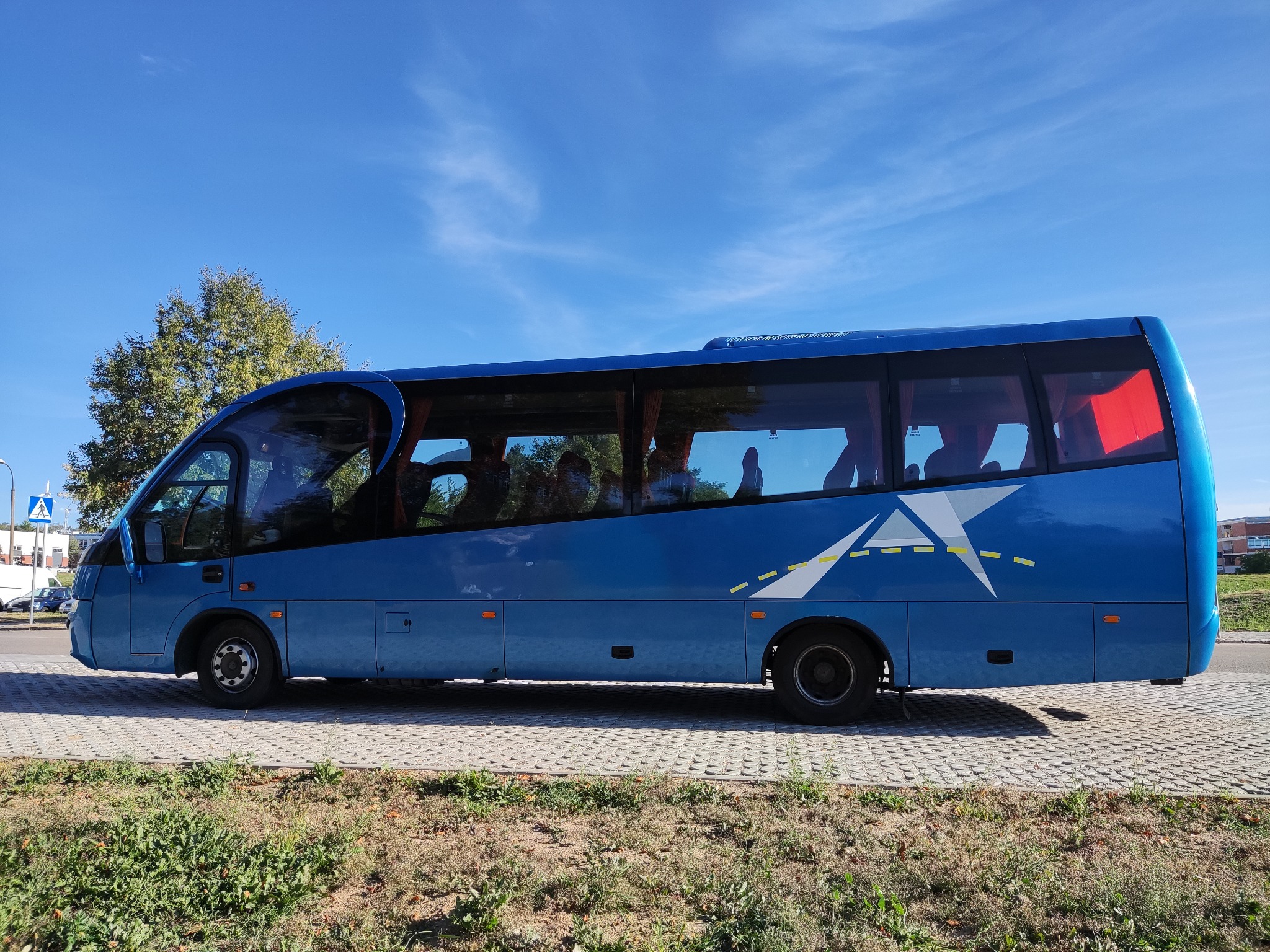 Niebieski autokar turystyczny z białym logo w kształcie gwiazdy i drogi, zaparkowany na brukowanym parkingu w słoneczny dzień.