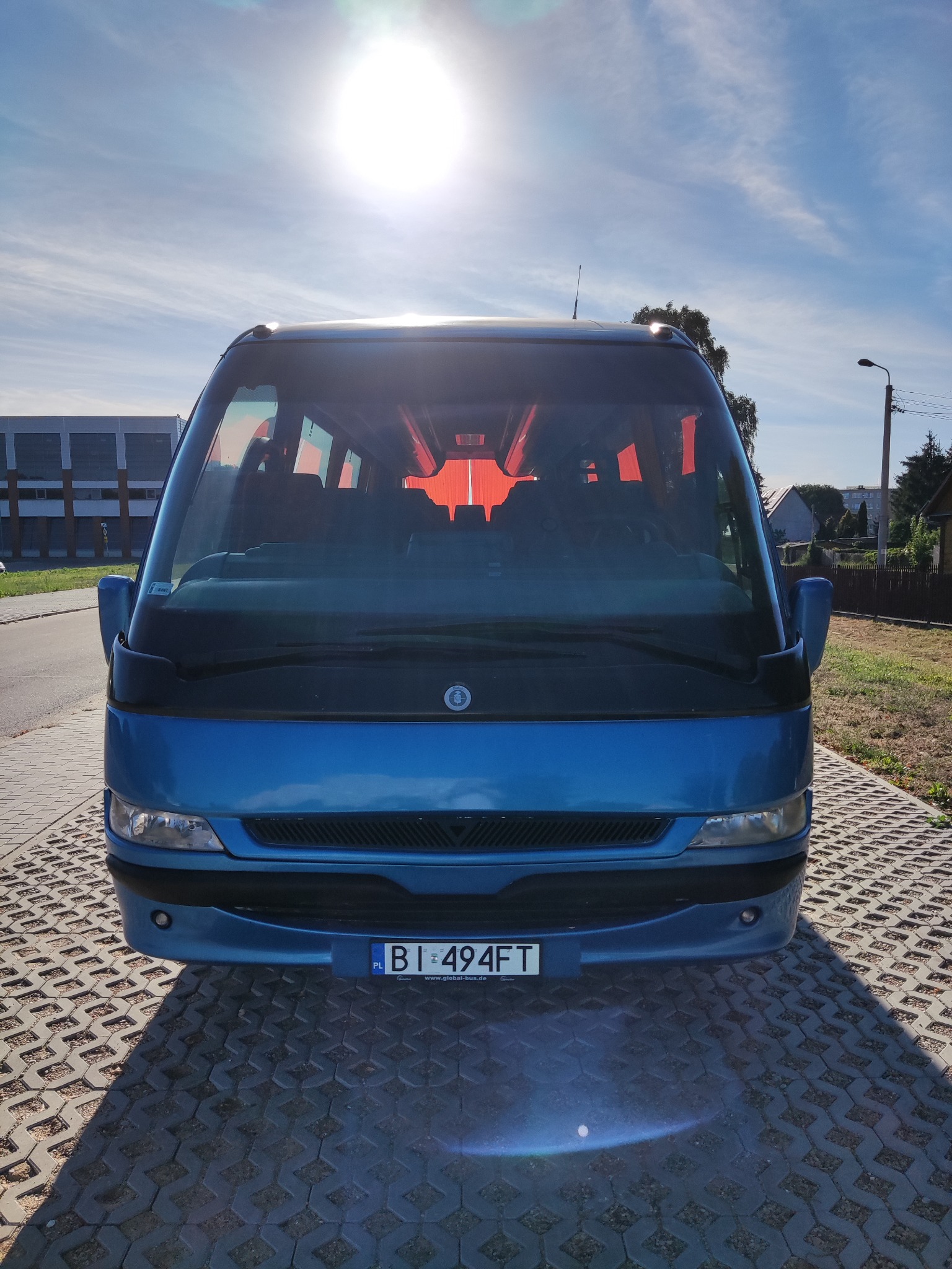 Niebieski bus z czerwonymi zasłonami w oknach, zaparkowany na kostce brukowej, z widoczną tablicą rejestracyjną BI 494FT.