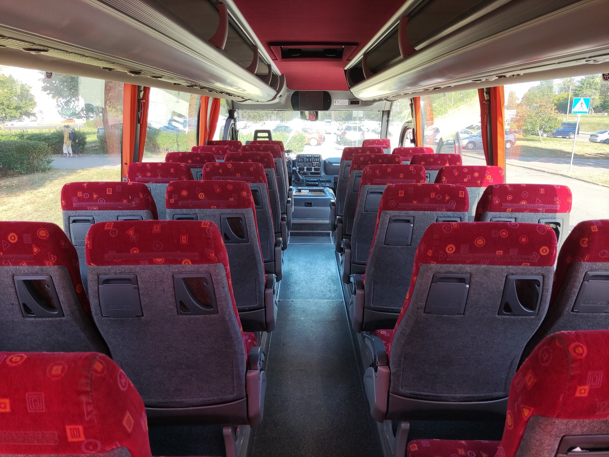 Wnętrze autobusu z czerwonymi siedzeniami z geometrycznym wzorem, widok na kokpit kierowcy i krajobraz za oknem z widocznym znakiem drogowym.