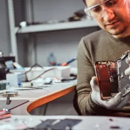 Technik w okularach ochronnych i rękawicach lateksowych naprawia rozłożony smartfon na warsztacie z narzędziami.