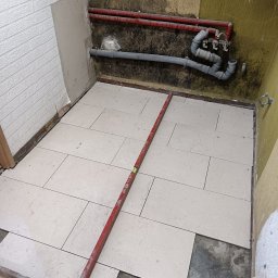 Stefan Palek - Podłoga w trakcie układania jasnych, marmurkowych płytek, widoczne rury i poziomica, ściany w surowym stanie przed wykończeniem.