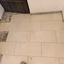 Stefan Palek - Układanie beżowych płytek podłogowych w korytarzu w Tarnowskich Górach, widoczne krzyżyki dystansowe i niedokończone fragmenty podłoża.