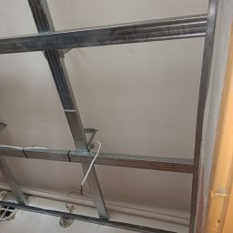 Stefan Palek - Metalowy stelaż sufitu podwieszanego z widocznymi przewodami elektrycznymi, zamocowany do surowego sufitu z widocznymi śladami wcześniejszych prac remontowych.