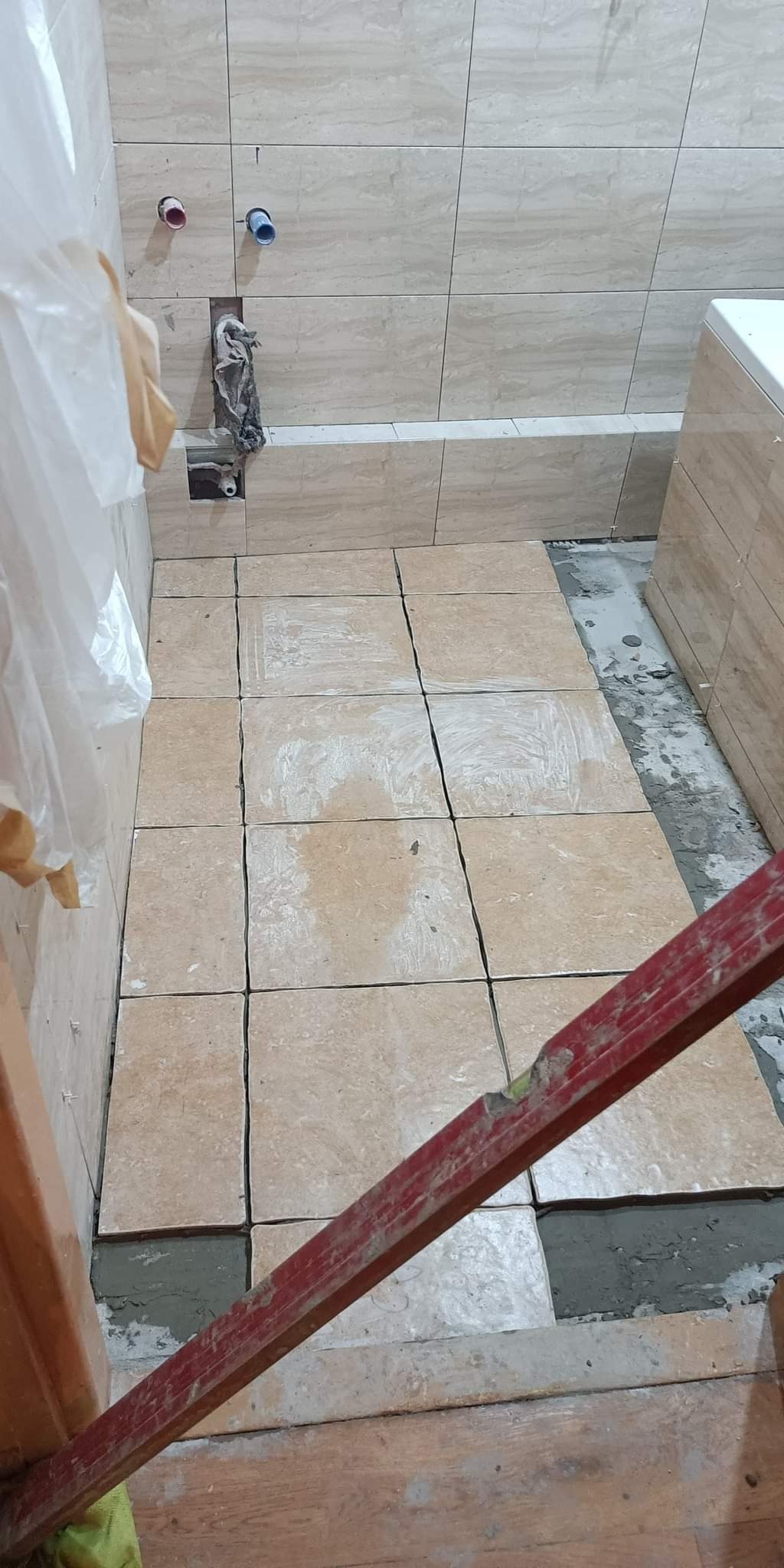 Podłoga w trakcie układania beżowych, kwadratowych płytek w łazience, widoczna poziomica i fragment drewnianej podłogi