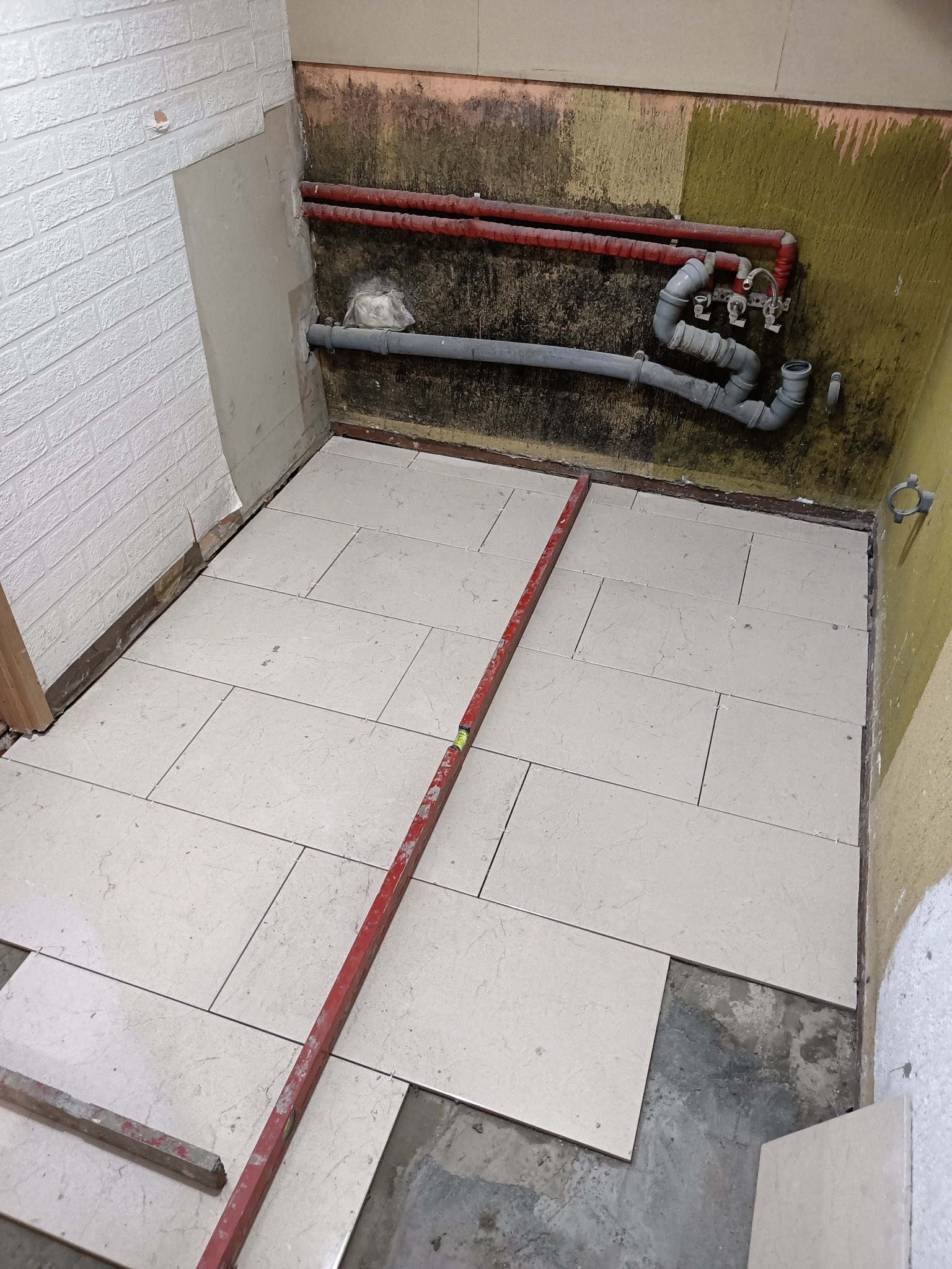 Układanie jasnych, marmurkowych płytek podłogowych w pomieszczeniu z instalacją hydrauliczną na ścianie, widoczny poziomica i fragment ściany z imitacją cegły.