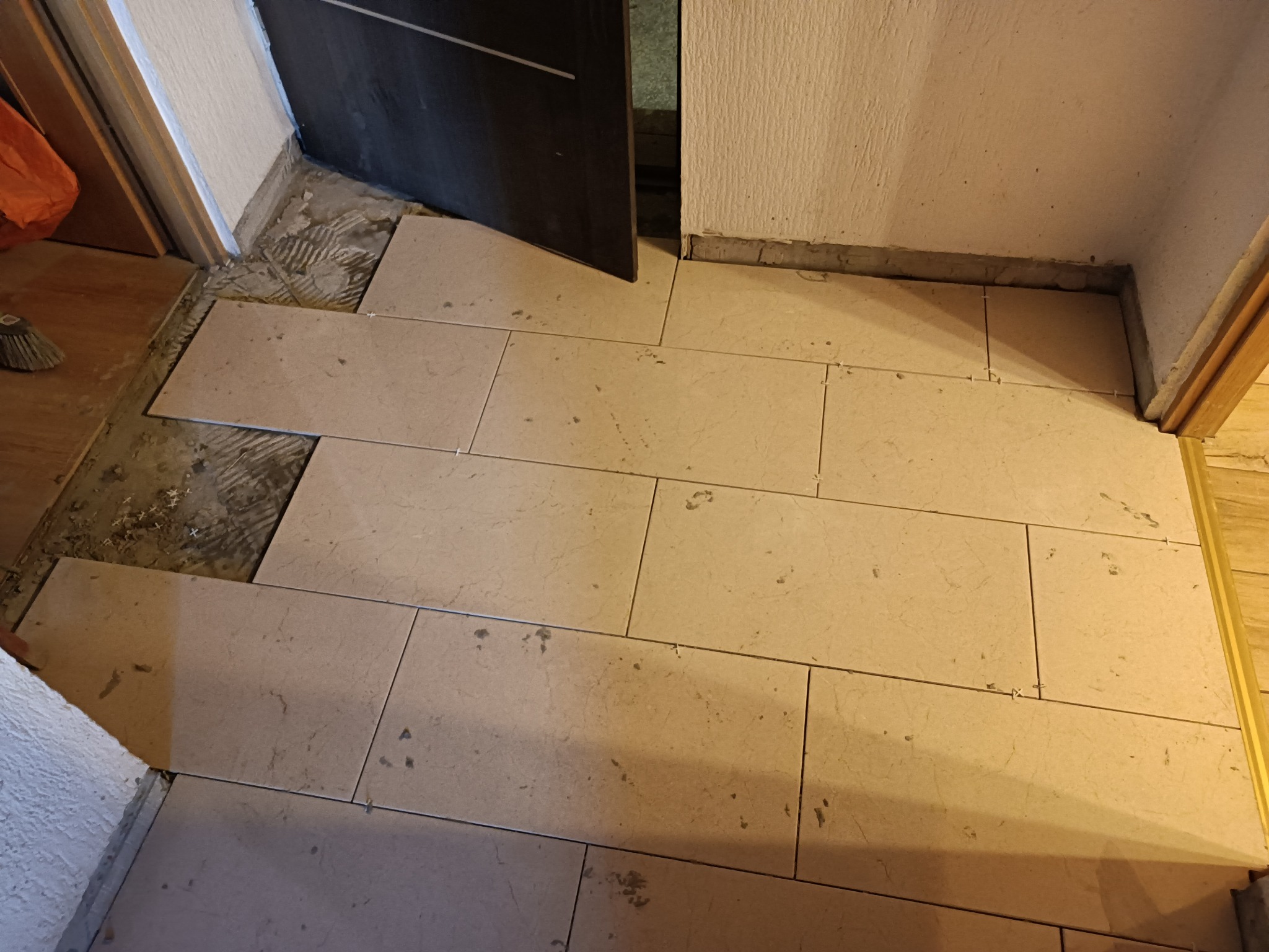 Układanie jasnych, prostokątnych płytek podłogowych w korytarzu, widoczne krzyżyki dystansowe między płytkami i fragment odsłoniętej wylewki podłogowej przy progu drzwi.