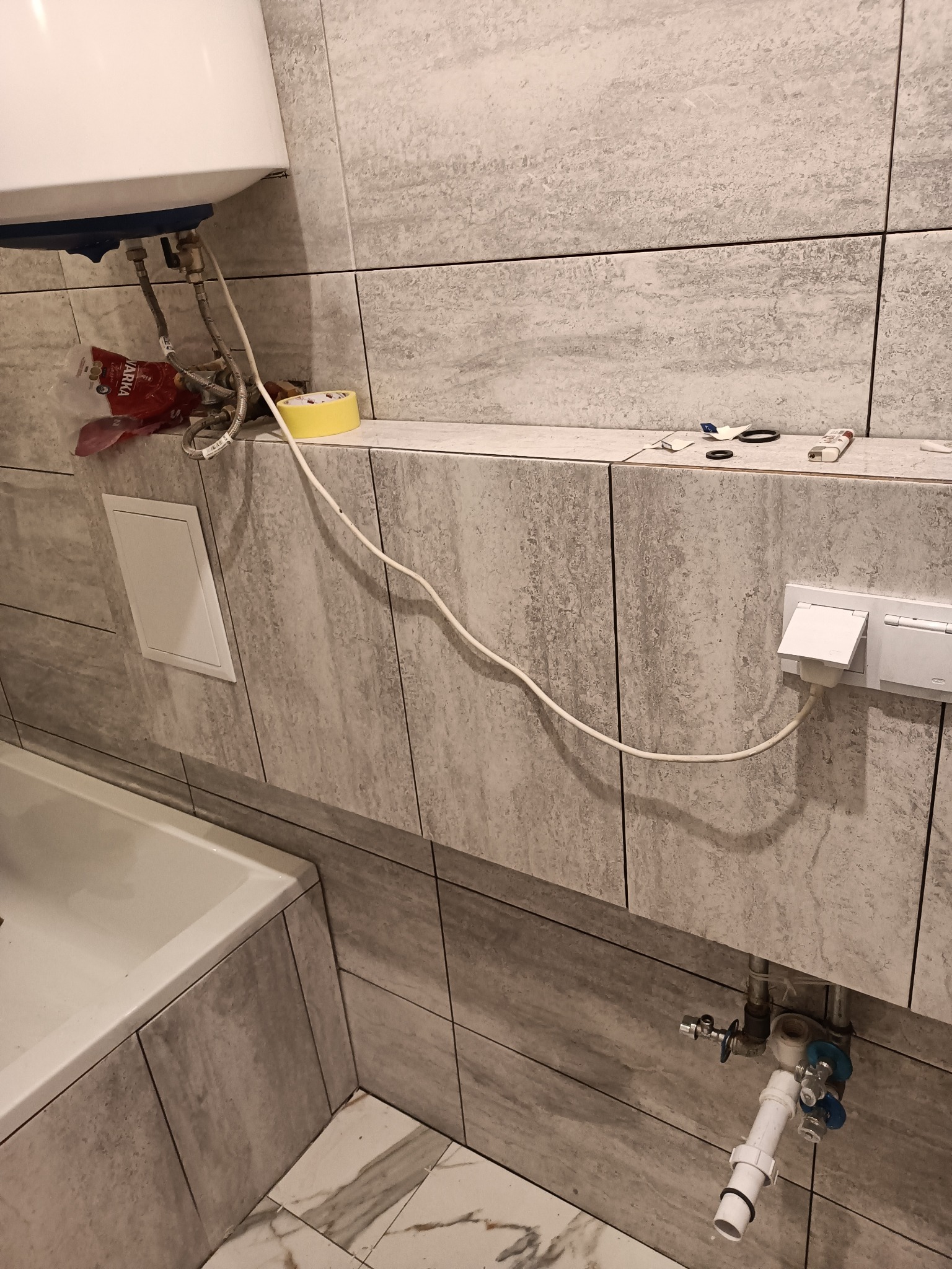 Fragment łazienki w trakcie prac: bojler podłączony do instalacji, widoczne płytki imitujące kamień, gniazdko elektryczne z podłączonym przewodem, akcesoria i narzędzia na półce.