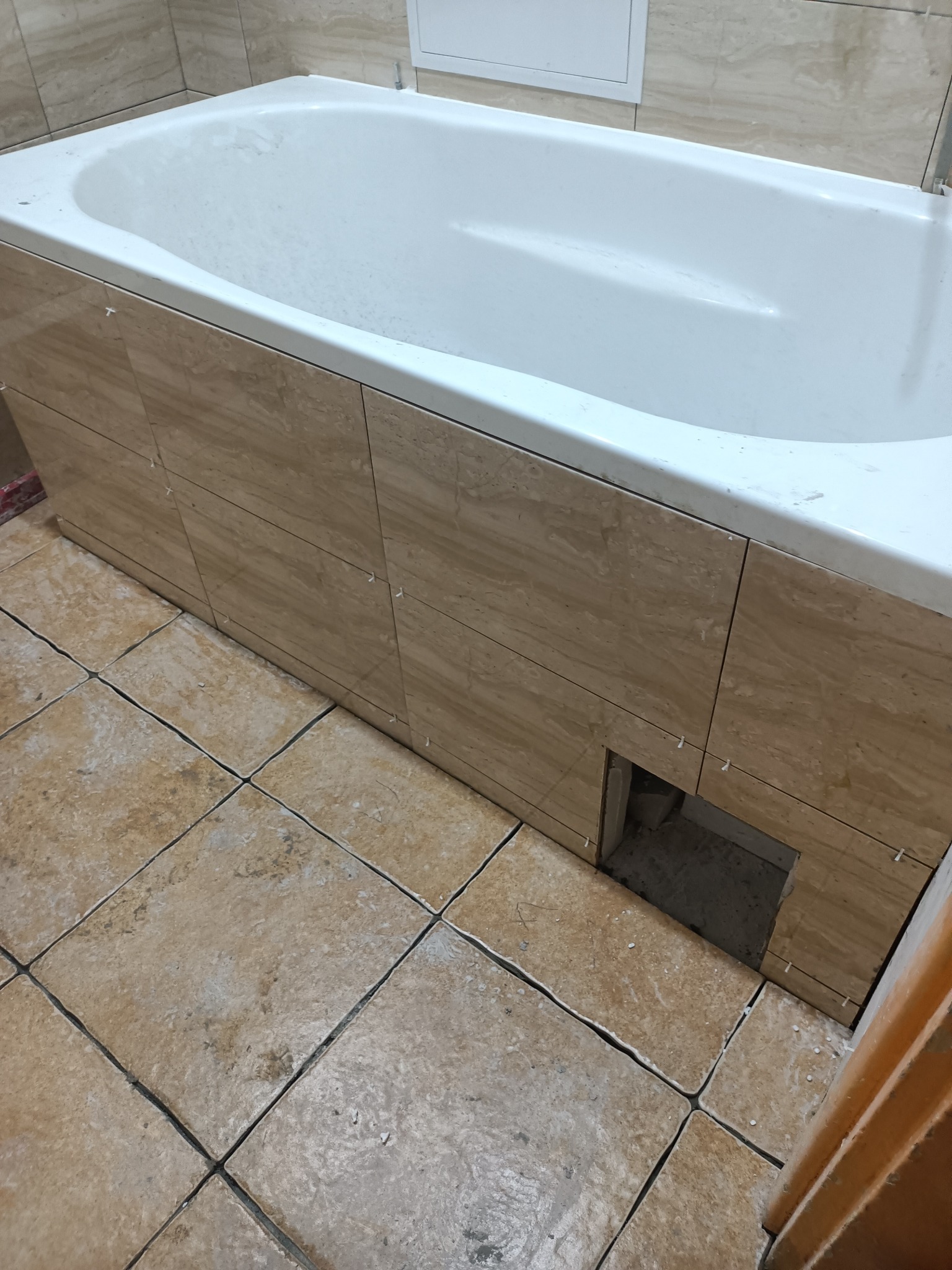 Biała wanna obłożona płytkami imitującymi drewno, z widocznymi krzyżykami dystansowymi, oraz podłoga wyłożona kwadratowymi płytkami w podobnym odcieniu beżu, otwór rewizyjny w obudowie wanny.