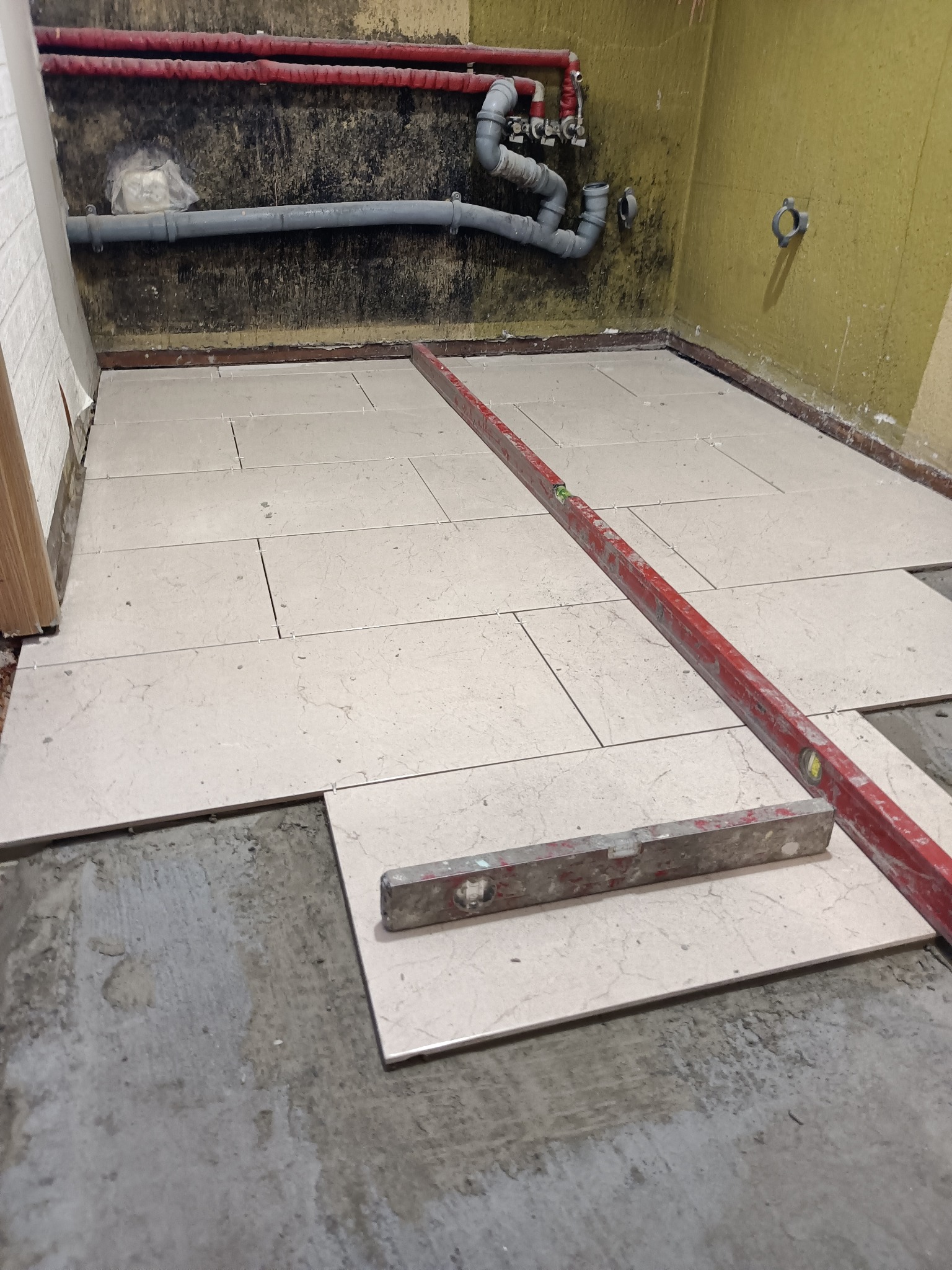Podłoga w trakcie układania płytek, widoczne poziomice, surowe ściany z rurami, częściowo ułożona jasna terakota.
