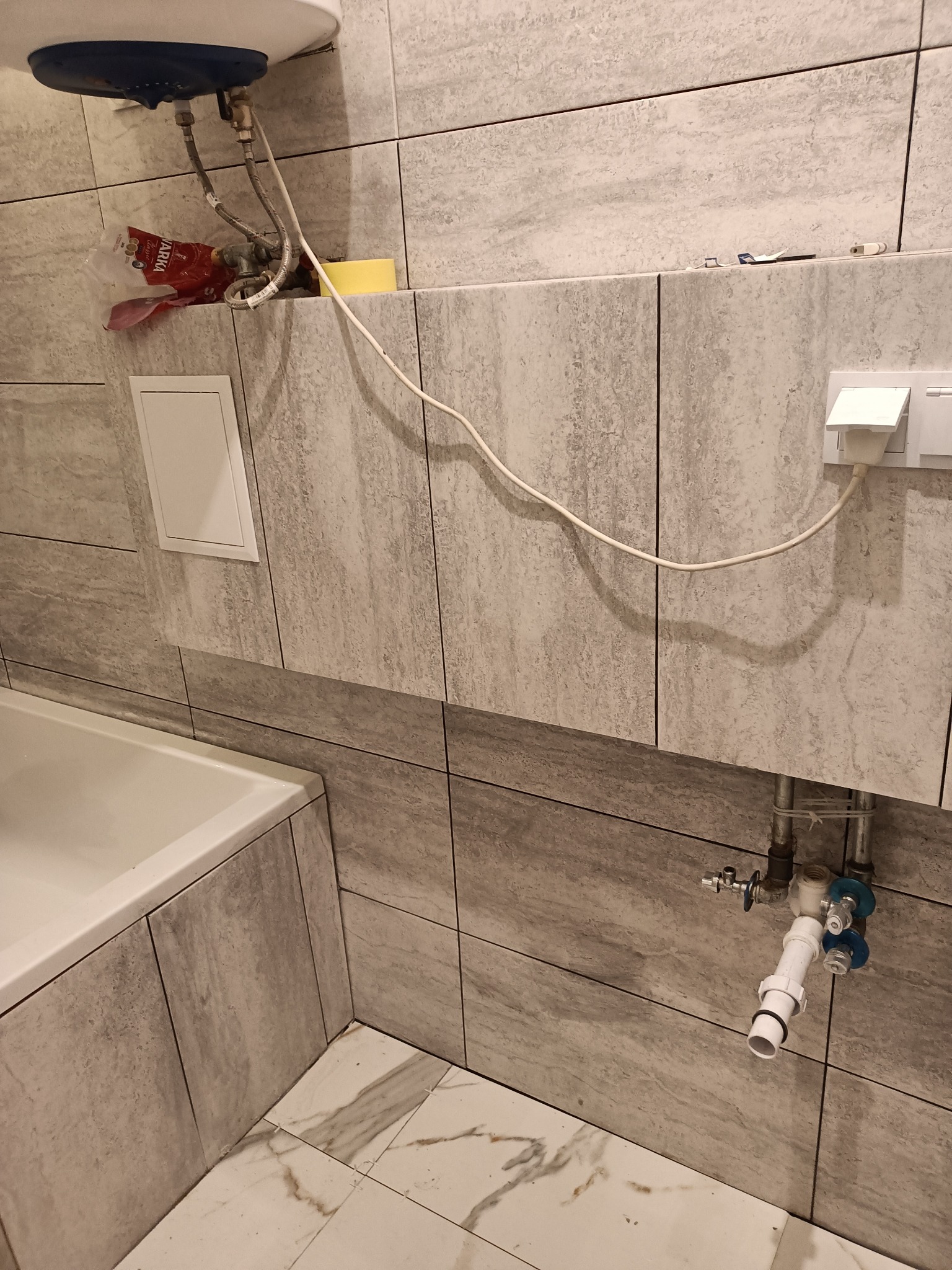 Szare płytki w łazience w trakcie montażu, widoczny bojler, instalacja hydrauliczna i elektryczna, gniazdka, fragment wanny i podłogi z marmurowym wzorem.