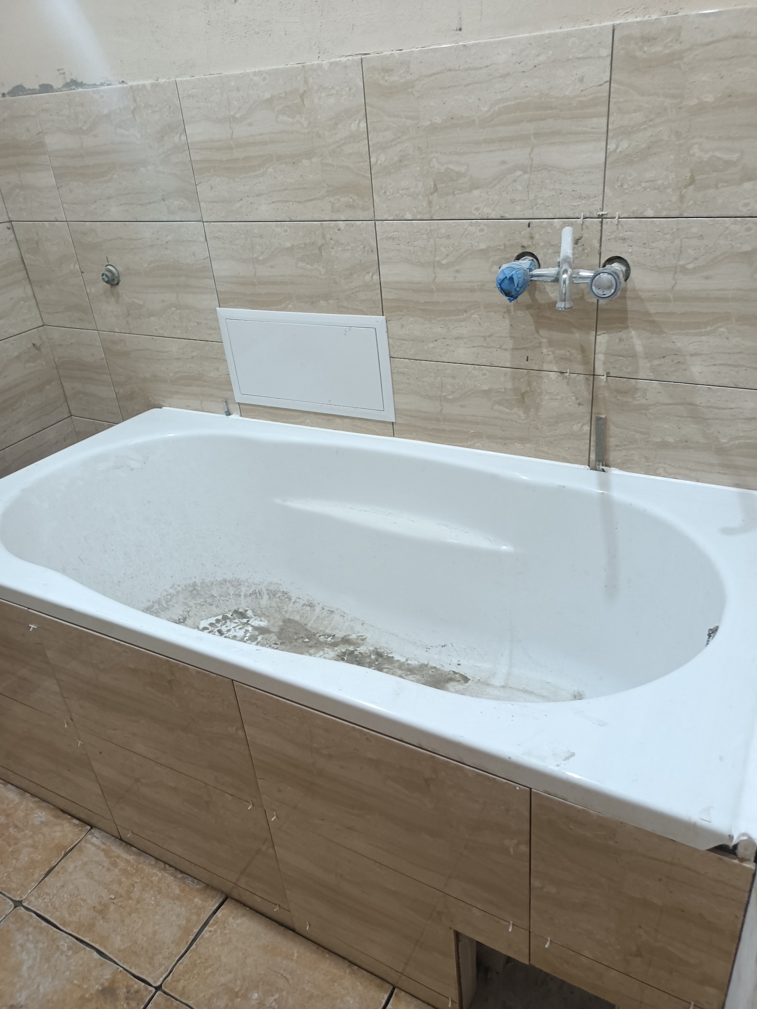 Wanna obudowana płytkami imitującymi marmur, widoczne krzyżyki dystansowe między płytkami, bateria zakryta niebieską folią, na dnie wanny pozostałości po pracach wykończeniowych.