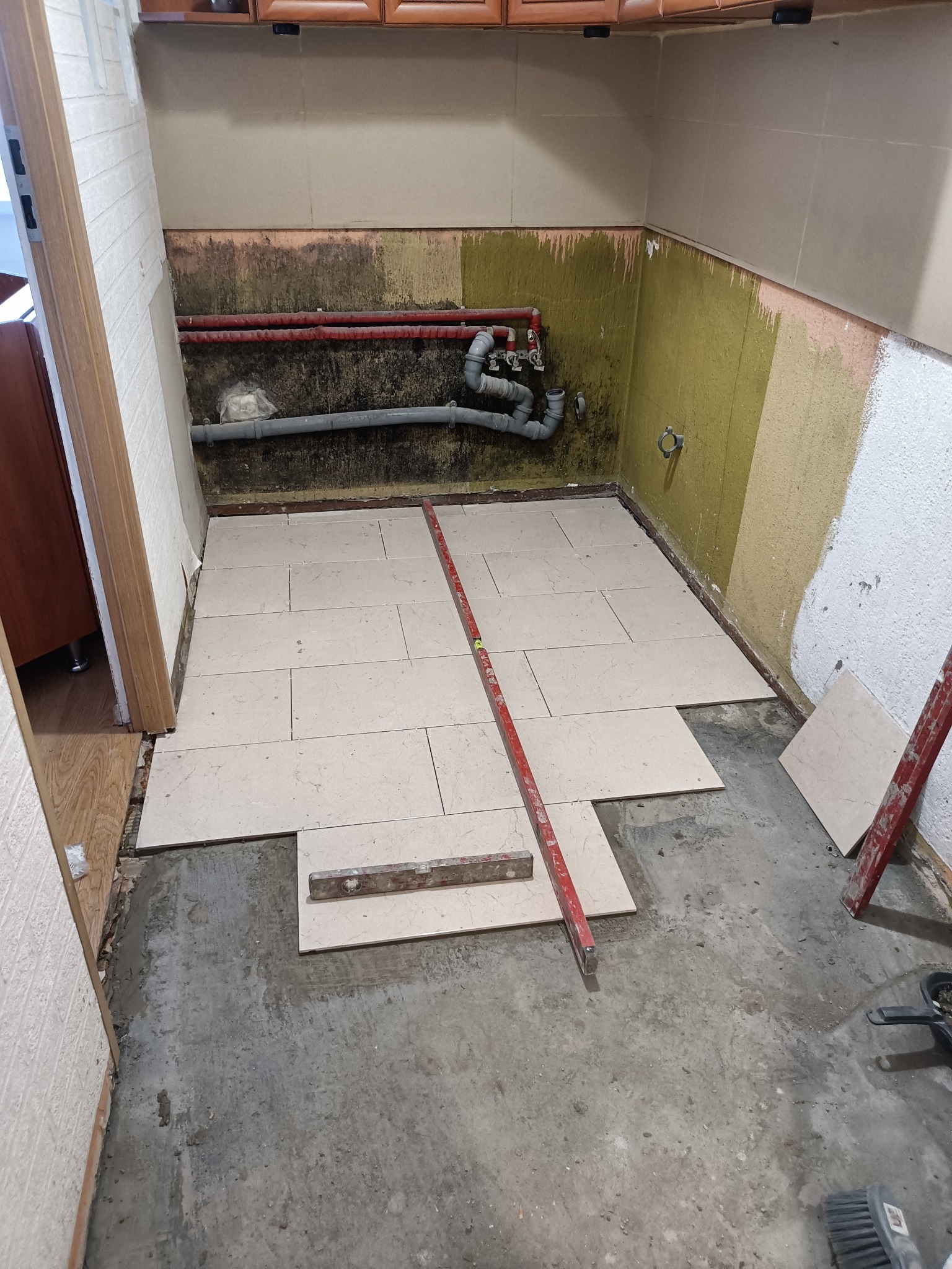 Układanie beżowych płytek podłogowych w małej kuchni w trakcie remontu, widoczne rury, poziomice i surowy beton na podłodze.