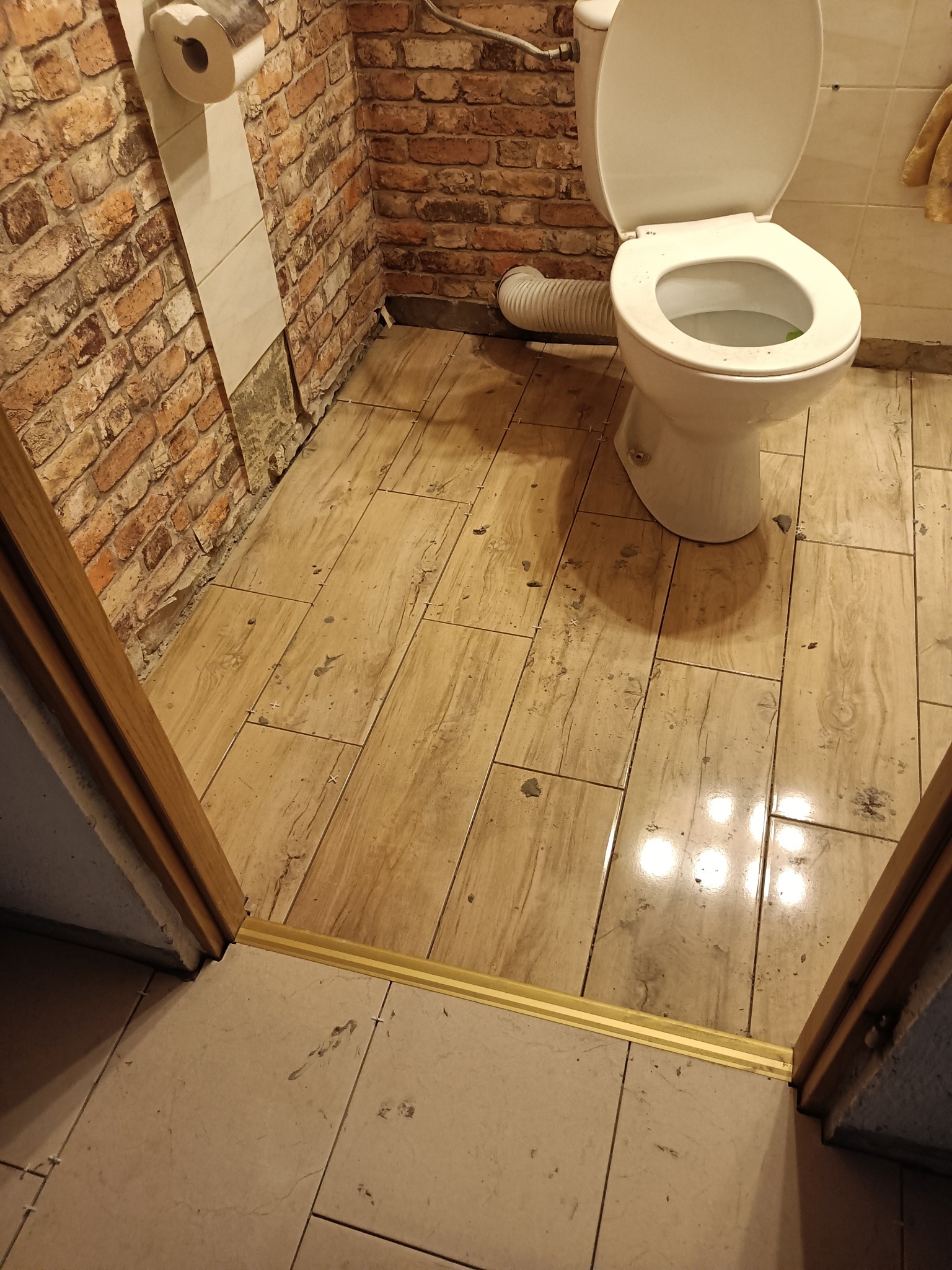 Ukończona łazienka z toaletą, brązowymi płytkami imitującymi cegłę na ścianie i drewnopodobnymi płytkami na podłodze, widoczne krzyżyki dystansowe między płytkami podłogowymi.
