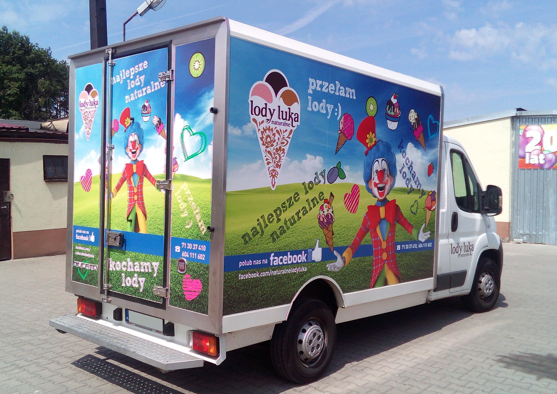 Oklejony samochód dostawczy z reklamą lodów Lody Luka z motywem klauna, logotypem i hasłami reklamowymi na tle łąki i nieba.