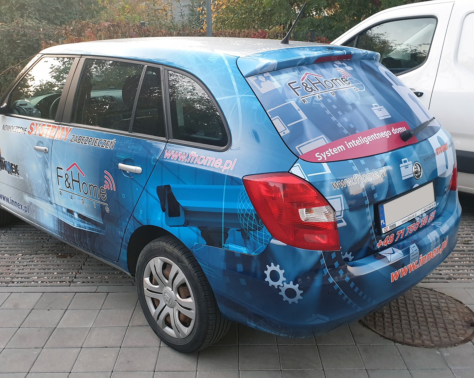 Niebieska Skoda Fabia oklejona folią z logo F&Home Radio i hasłem 'System inteligentnego domu', zaparkowana na chodniku z kostki.